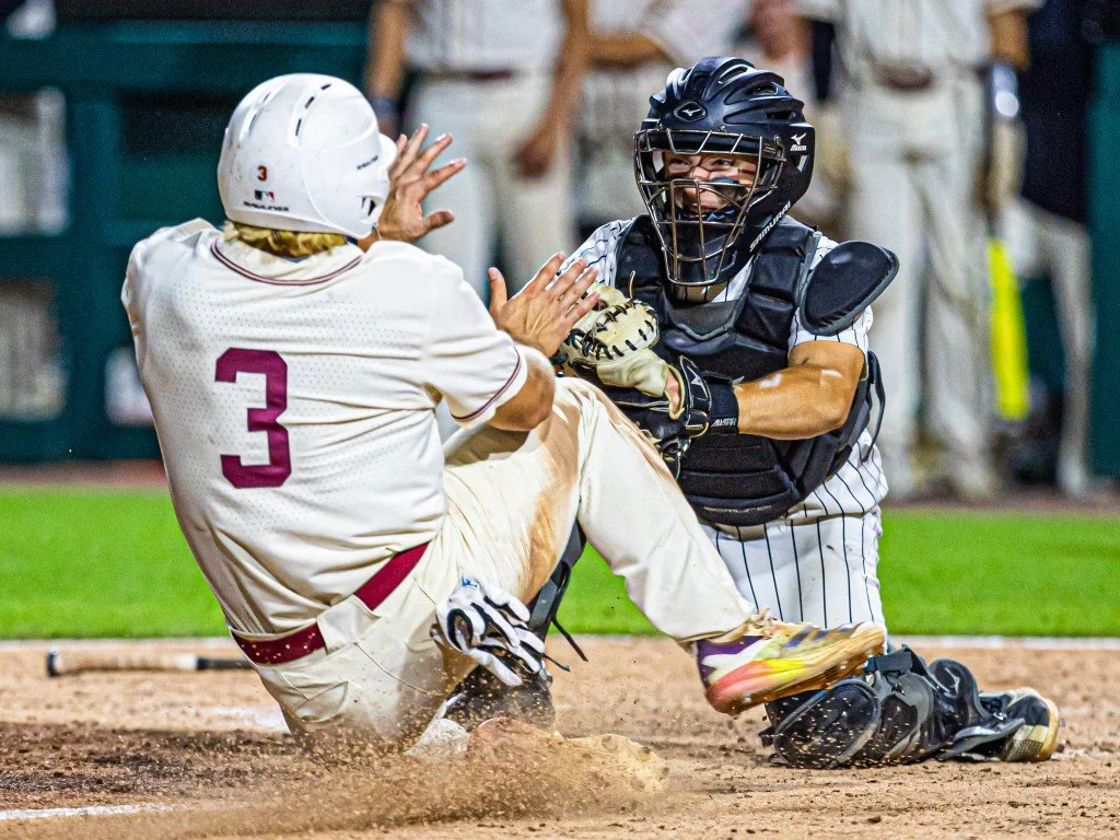 A baseball player in a white uniform with the number 3 slides into home plate, being tagged by a catcher in black protective gear. The player is wearing a white helmet, and the catcher is wearing a black helmet and chest protector. The scene is happe