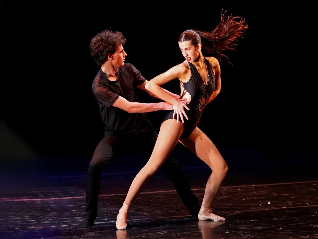A male and female ballet dancers perform a dance on stage, with the male dancer holding the female dancer's waist and the female dancer striking a dramatic pose with her hair flowing.