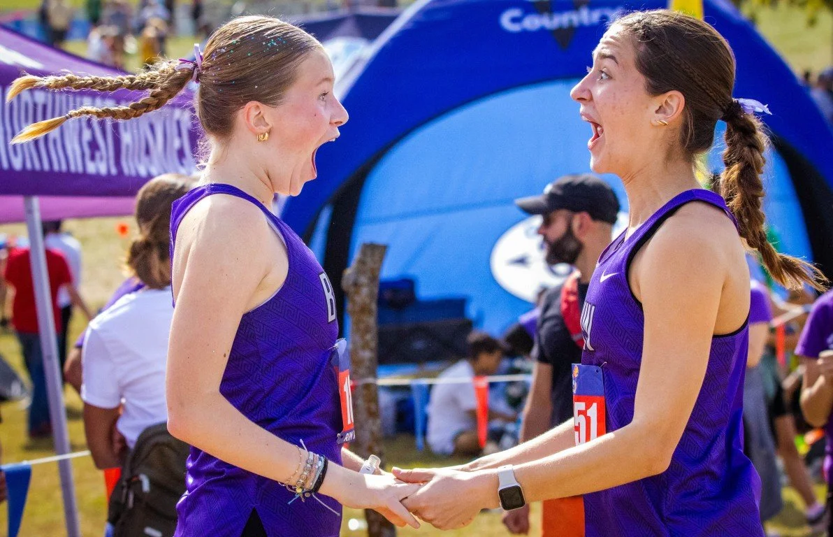 Two female runners in purple athletic uniforms holding hands and smiling during a race, with a blue sponsor arch and other runners in the background.