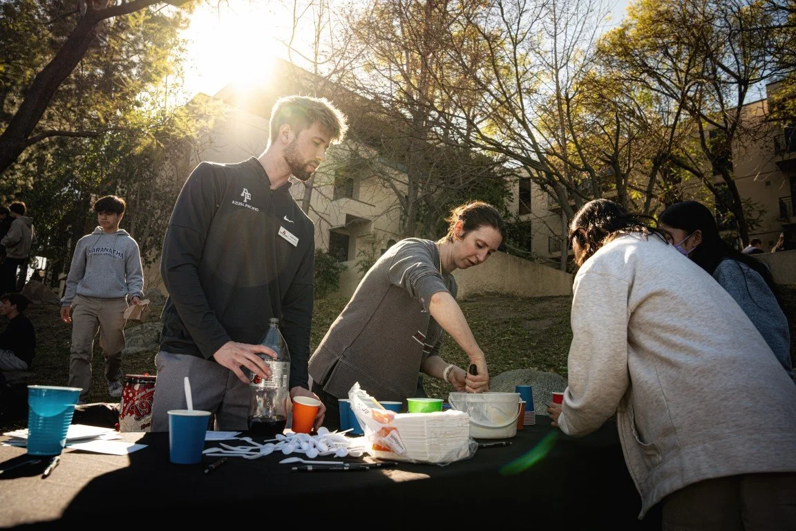 People gathered around a table outdoors, serving and preparing food, with trees and buildings in the background and sunlight shining through the trees.