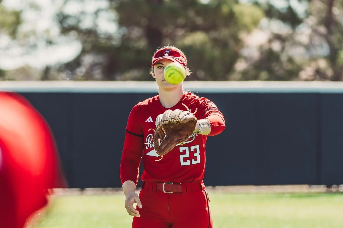 A female softball player in a red uniform with the number 23 on her jersey catches a yellow softball with her glove during a game.