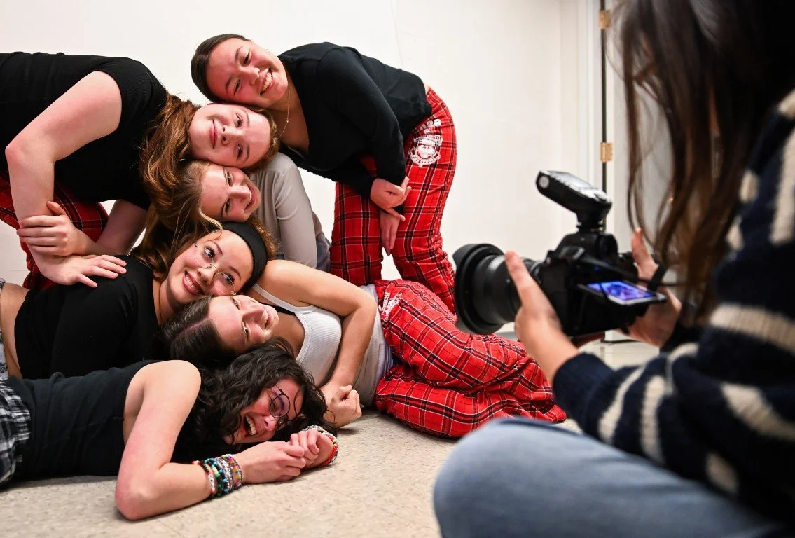 Group of six young women lying and sitting on the floor, posing together for a photo taken by a person holding a DSLR camera.