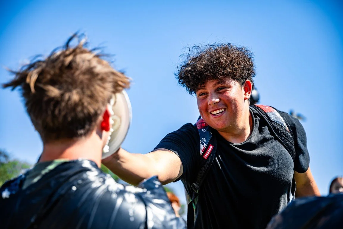 A young man with curly hair smiling and extending his hand to shake hands with another person whose back is turned to the camera, outdoors on a sunny day.