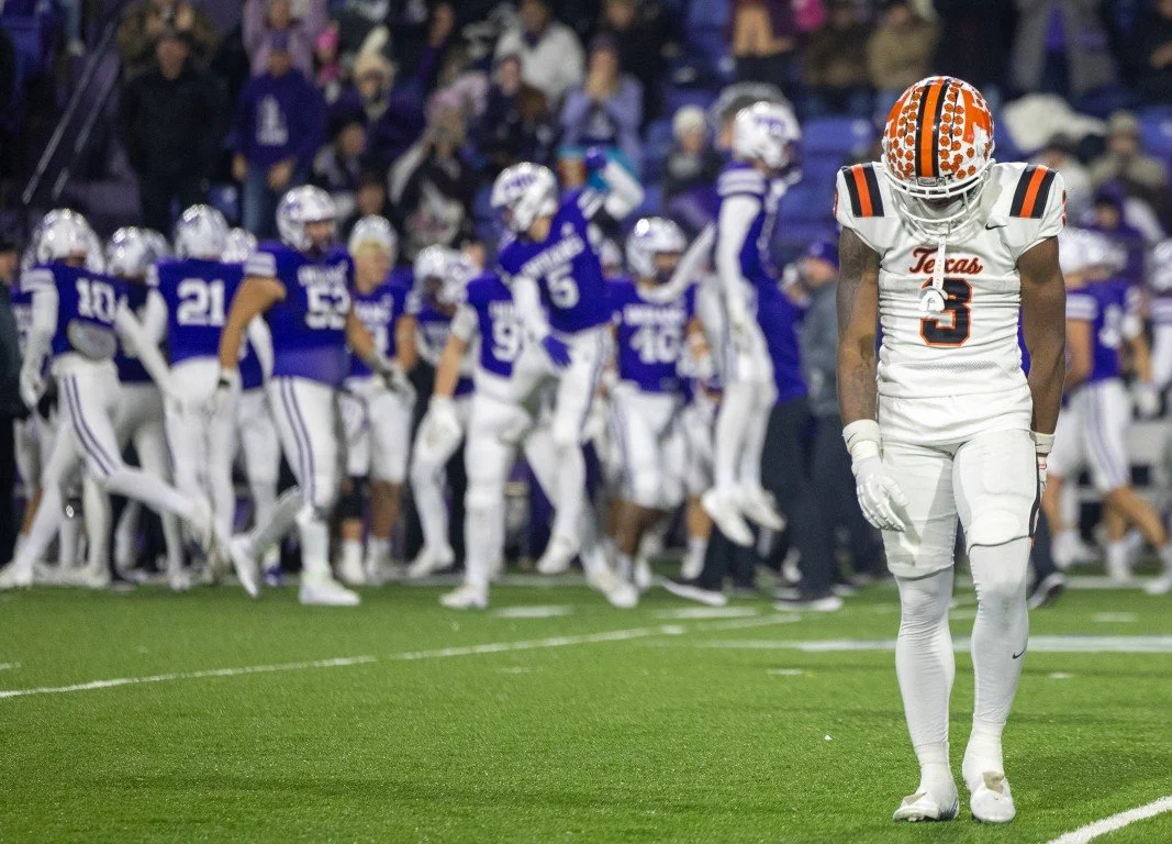 A football player in a white uniform with orange and black helmet, standing alone on the field, while his team in blue uniforms is in the background.