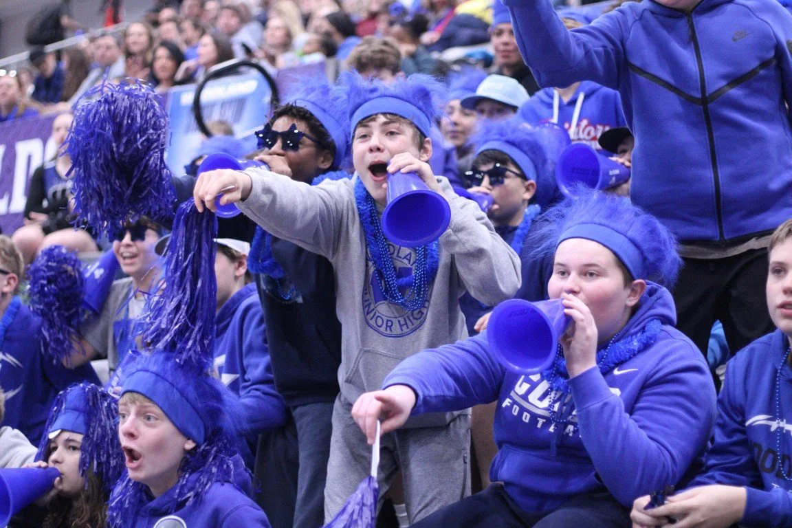 Group of children dressed in blue, cheering and using megaphones at a sports event with many spectators in the background.