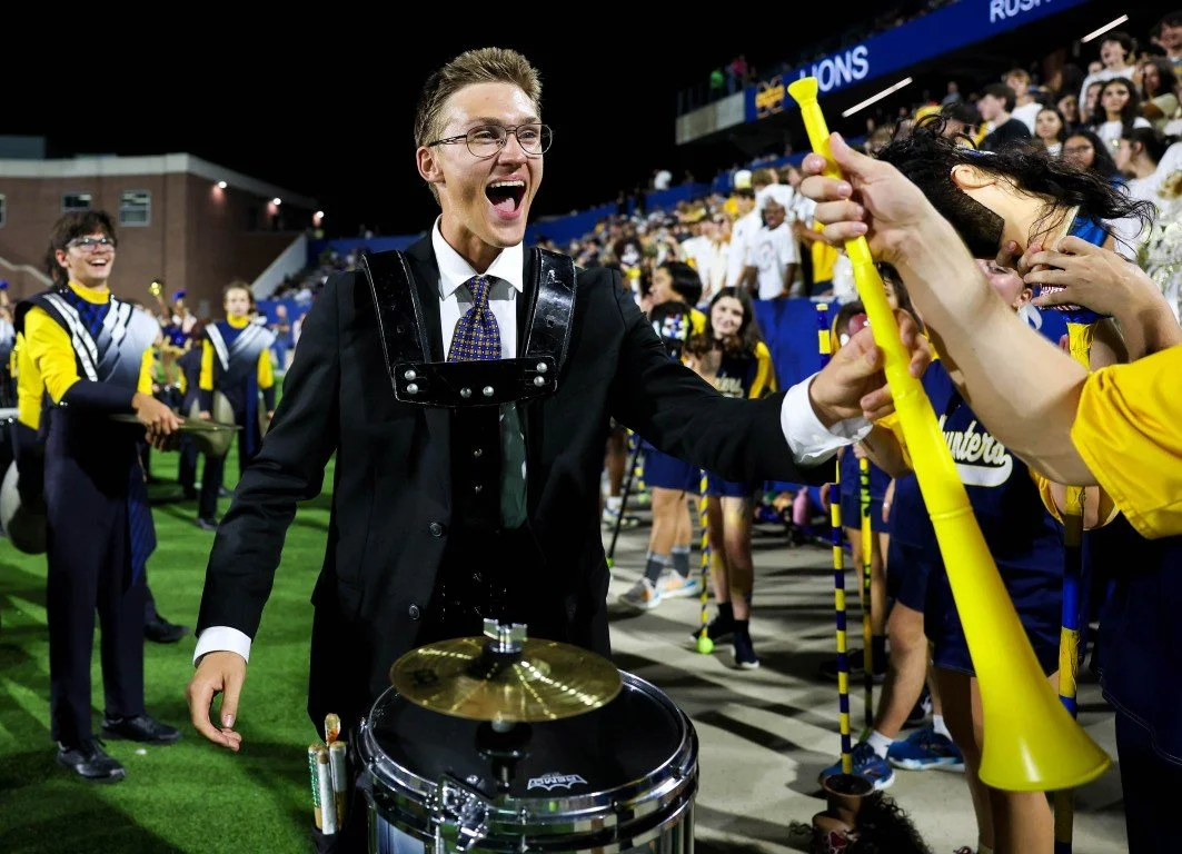 Celebrating fans and band members at a sporting event, with a woman in a suit and glasses smiling and interacting with spectators near a drum set.