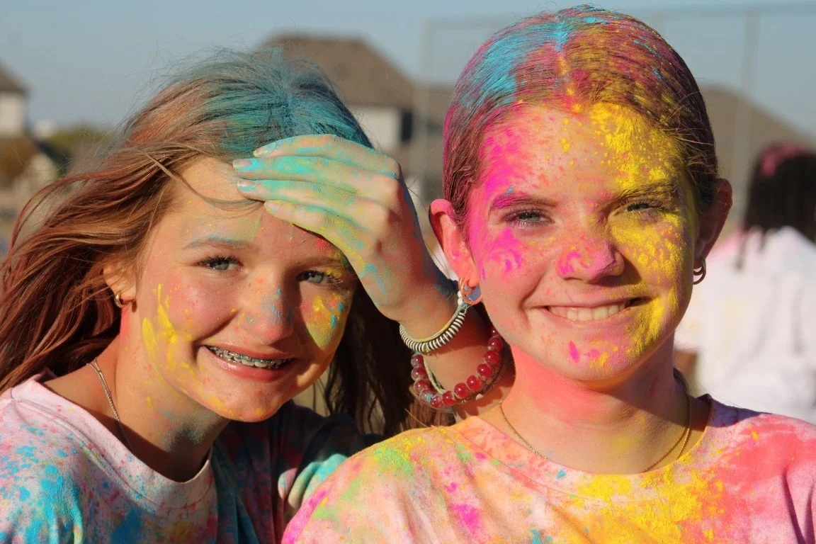 Two young girls covered in colorful powder at a celebration or festival, smiling outdoors.
