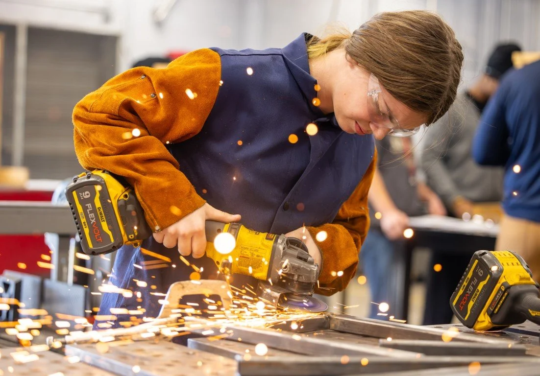 A woman working with a power tool in a workshop, wearing safety glasses and a blue and orange apron.