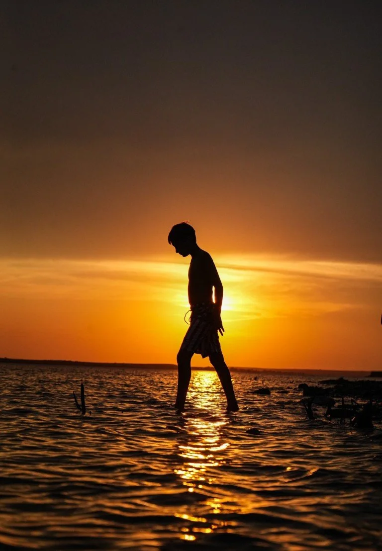 A silhouette of a boy walking in shallow water during sunset.