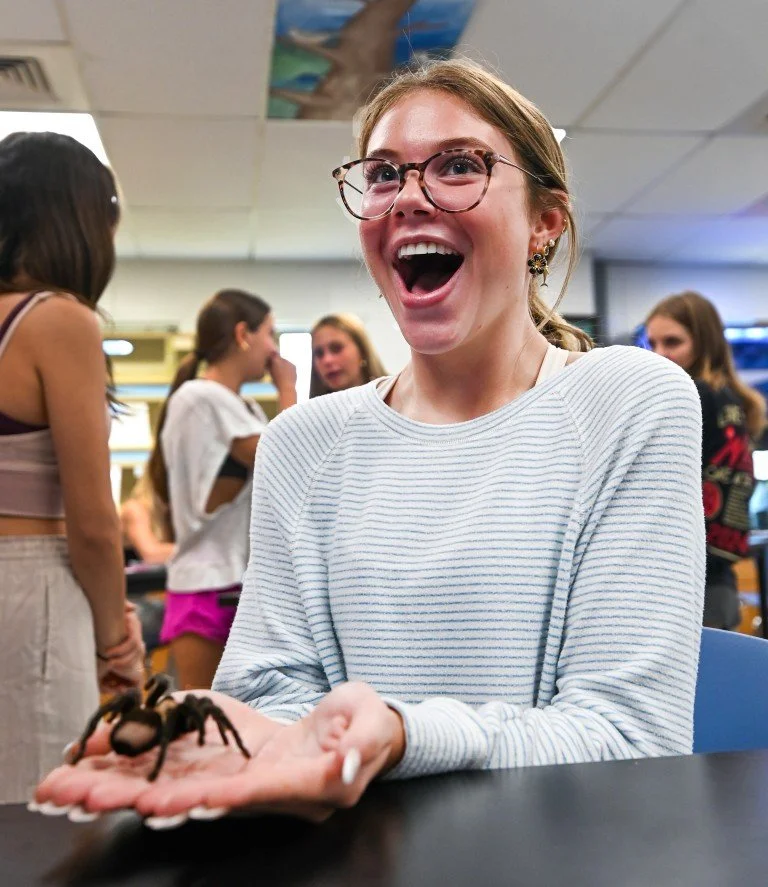 A young woman with glasses smiling and holding a large spider in her hand in a room with other people in the background.