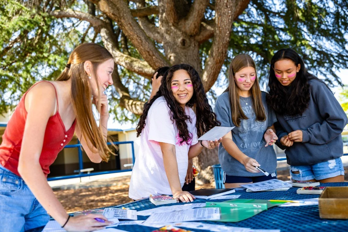 Four teenage girls with pink face paint on their cheeks, standing around a table outdoors under a large tree, looking at papers and smiling.