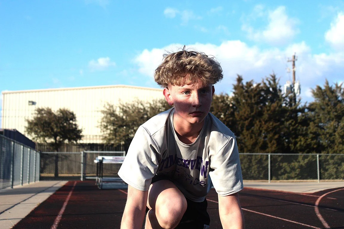 A young boy with curly blonde hair crouching on a tennis court during daytime, wearing a gray T-shirt and black shorts, with an intense expression, sunlit scene with a blue sky and trees in the background.