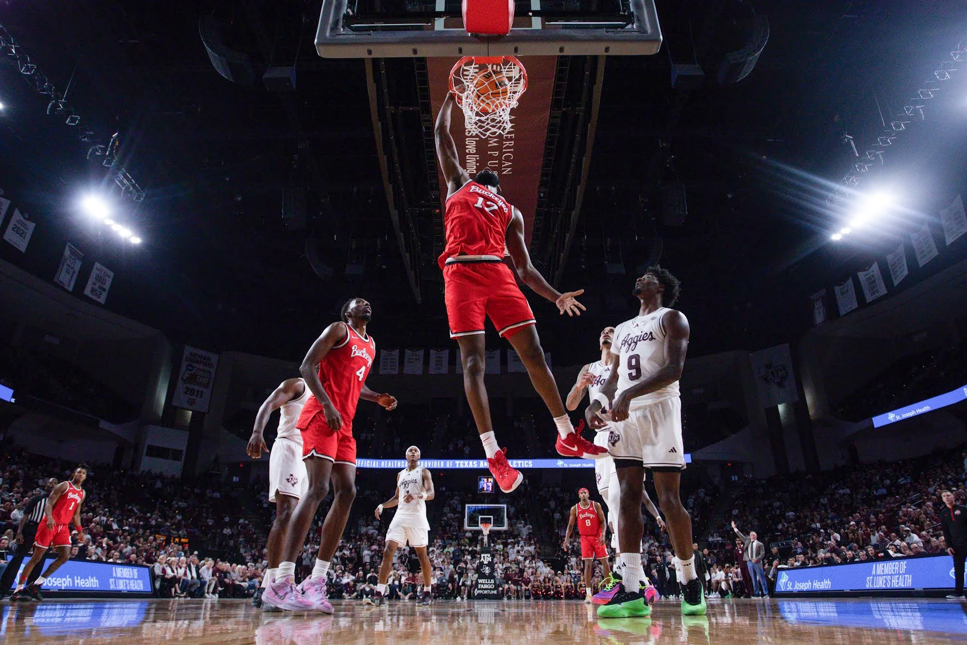 Basketball game with player in red jersey jumping to make a dunk, surrounded by players in white jerseys, in a packed indoor arena.