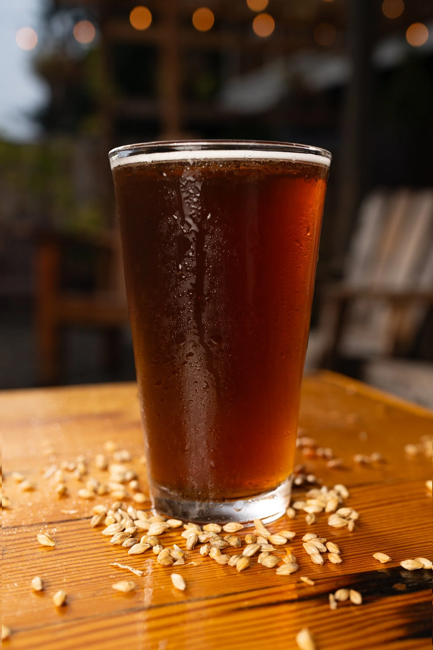 A tall glass of dark soda with condensation on the outside, placed on a wooden surface with scattered grains or seeds around it, and a blurred background with warm lights.