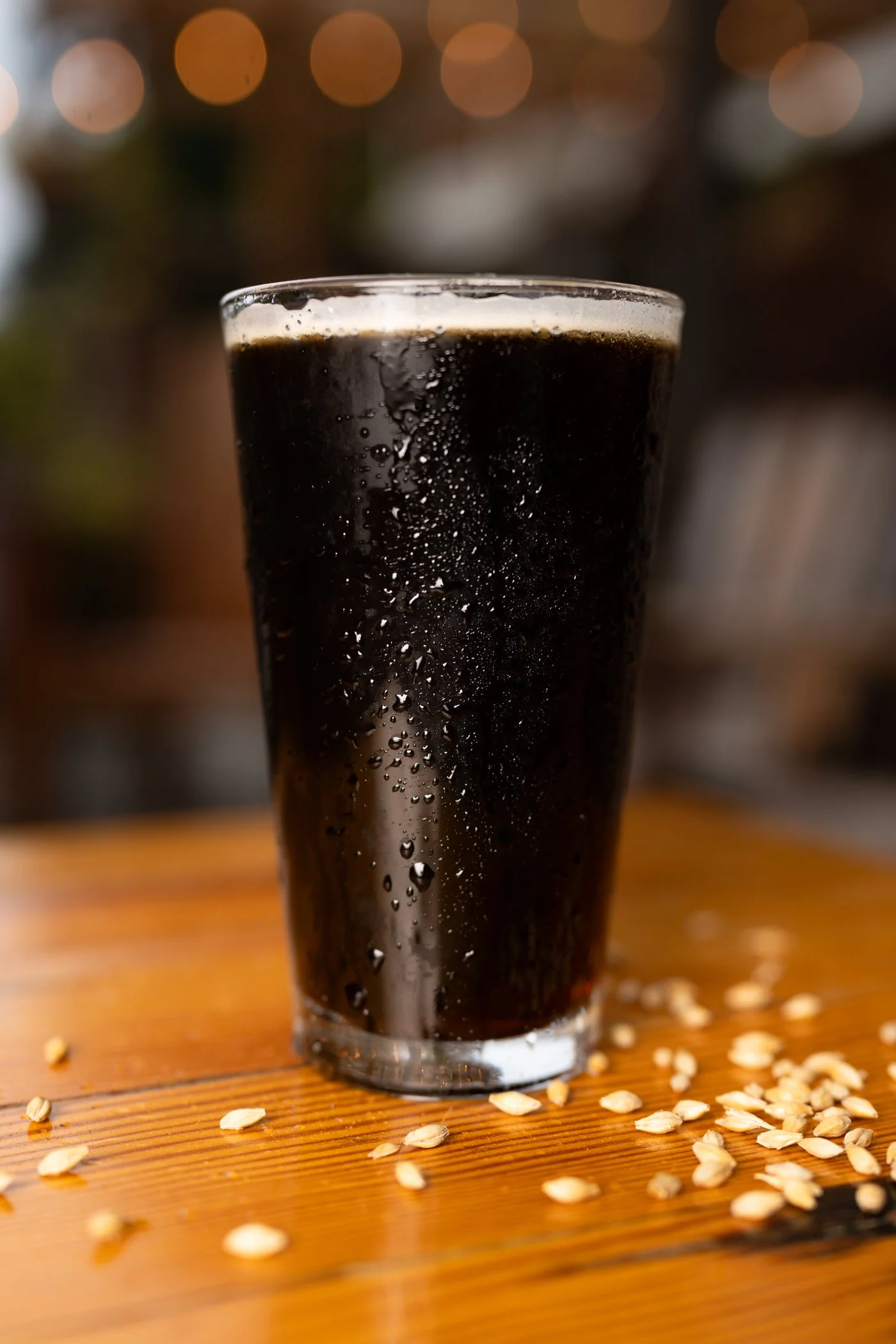 A glass of dark soda with condensation, placed on a wooden surface with scattered oats around it, with a blurred bokeh background.