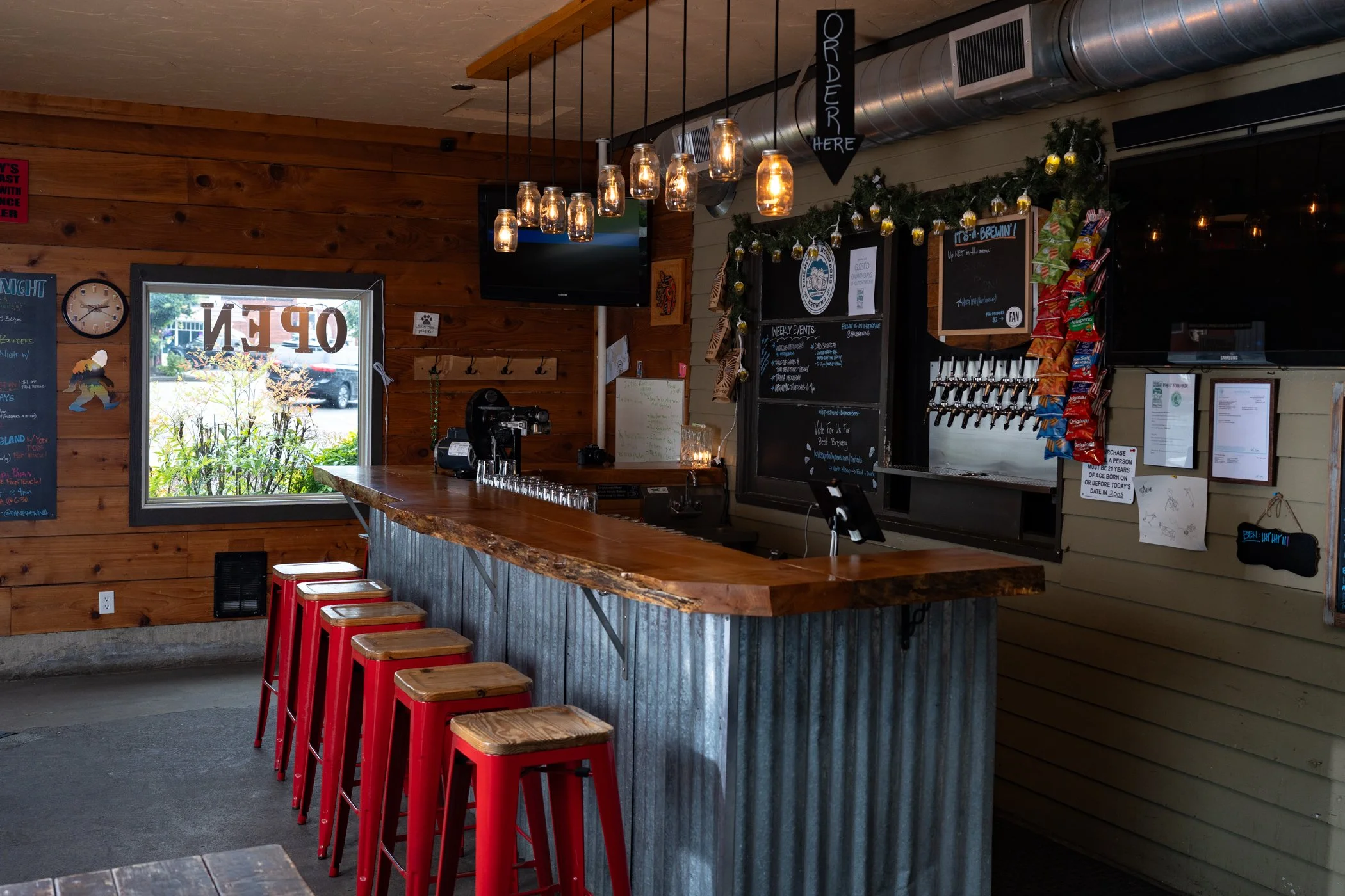 Interior of a restaurant or bar with wooden walls and a wooden bar counter. Red bar stools are lined up at the counter. Hanging light fixtures made of jars with warm lighting. Signs, a chalkboard menu, and decorative garlands are visible behind the counter. A window with an 'OPEN' sign, and a flat-screen TV are also present.