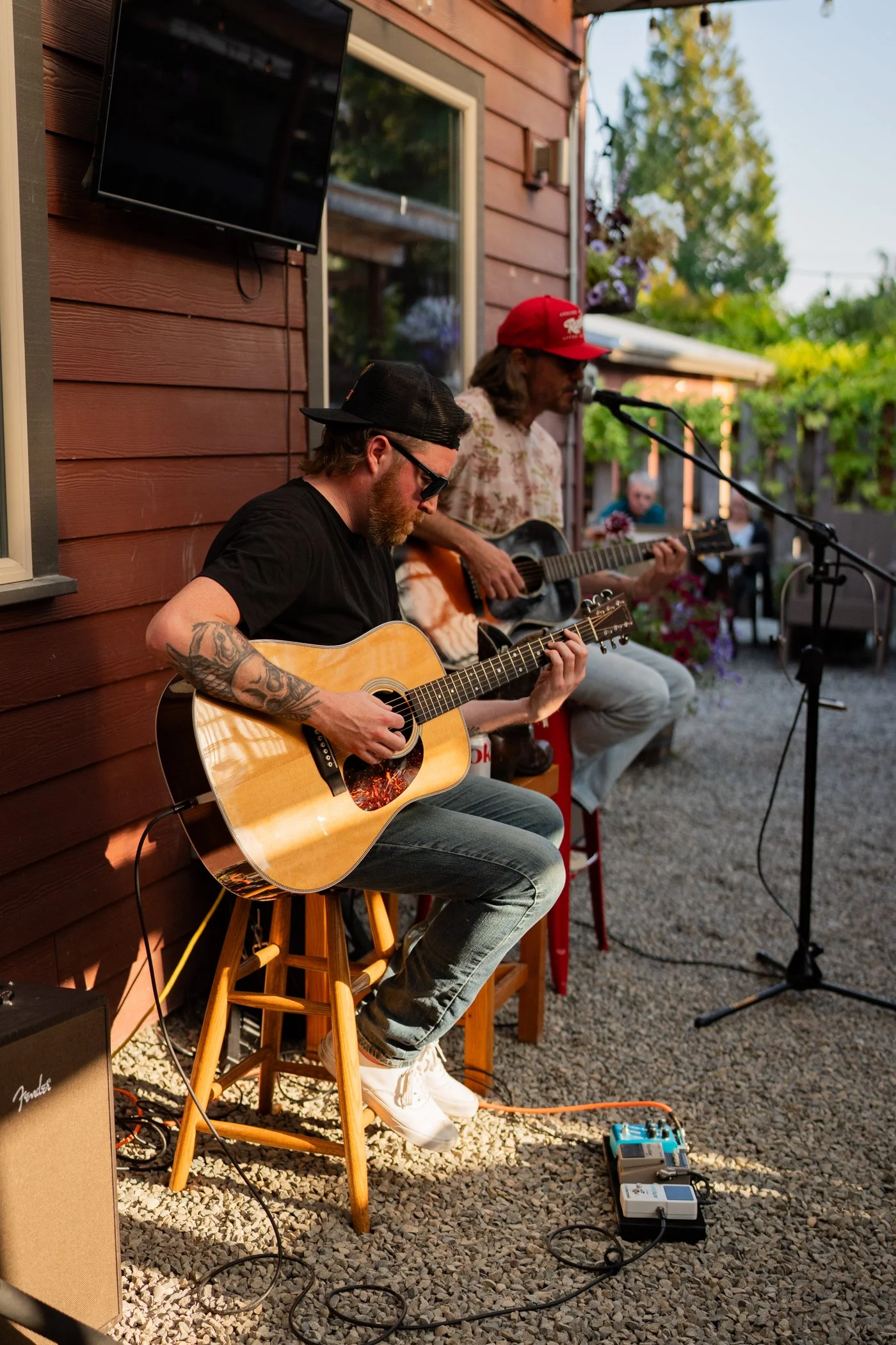 Two musicians performing outdoors on a gravel surface. One is playing an acoustic guitar, wearing a black cap, glasses, a black t-shirt, and has tattoos on his arm. The other is singing into a microphone and playing an electric guitar, wearing a red cap, sunglasses, and a floral shirt. There are amplifiers, pedals, and a monitor speaker nearby. The setting appears to be a backyard patio with a wooden house and greenery.