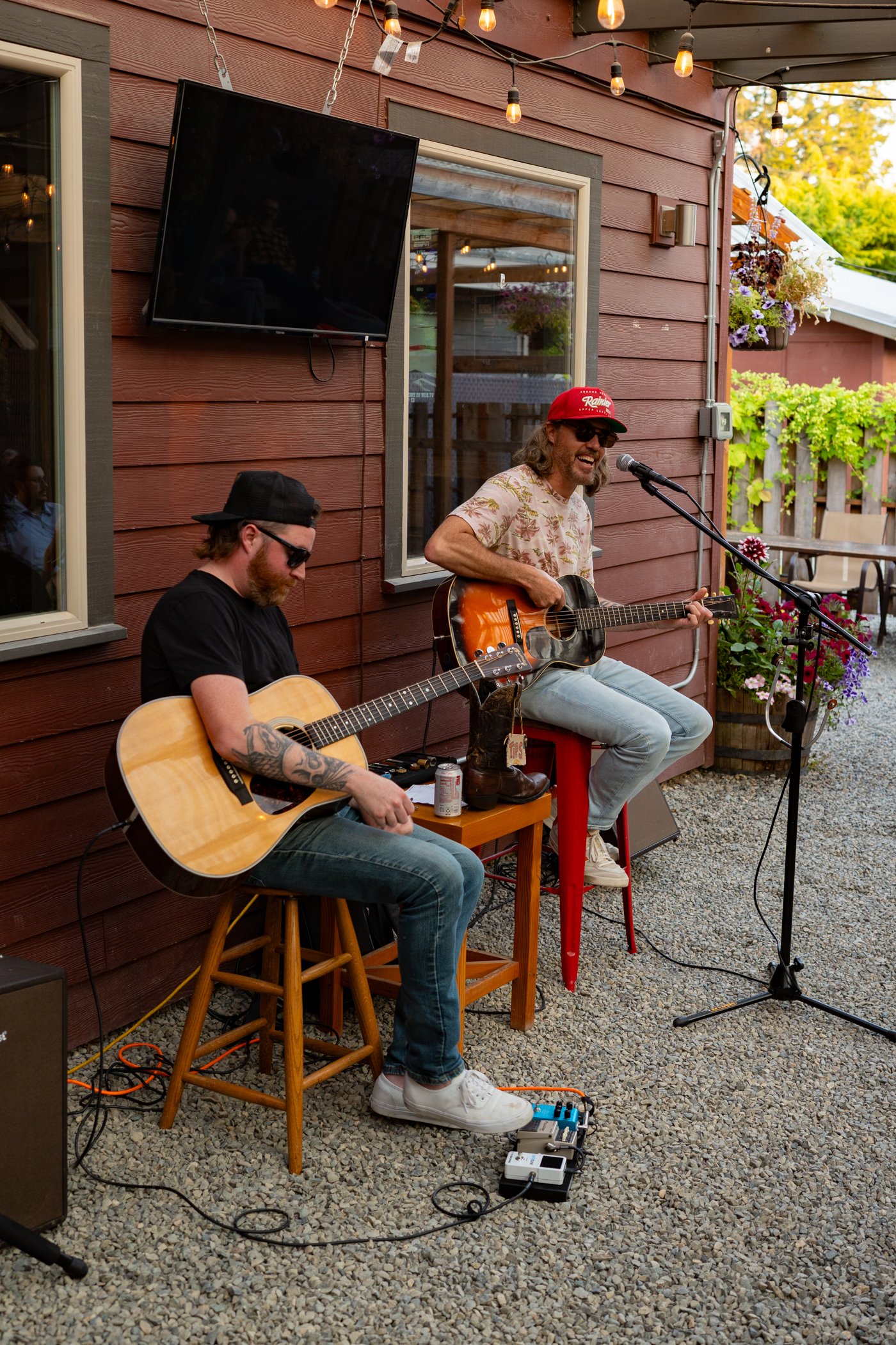 Two musicians performing outdoors at a patio with a red wooden wall, string lights, and flower decorations. One is playing acoustic guitar and the other is singing into a microphone while playing an electric guitar.