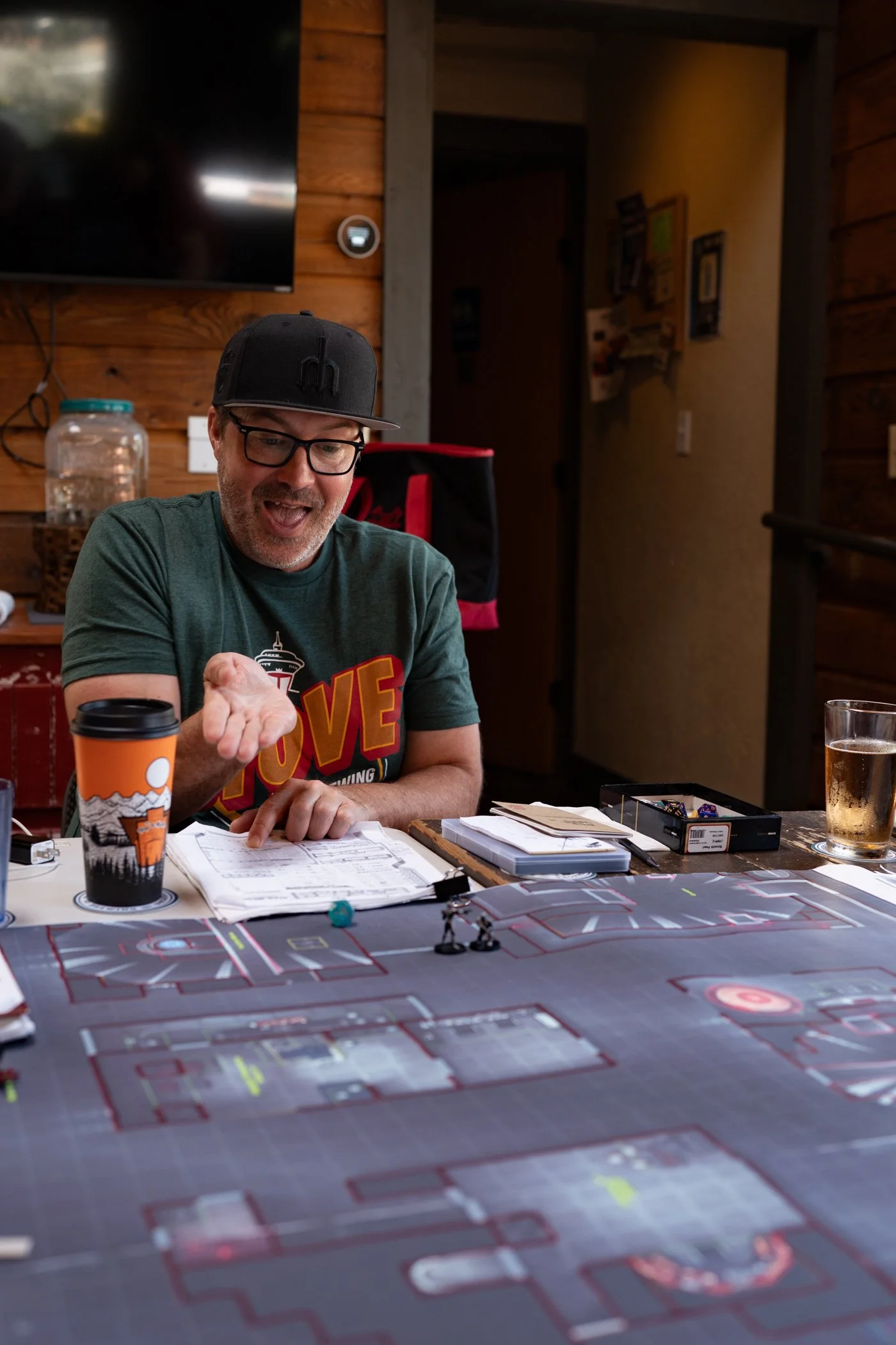 A man wearing glasses and a black cap sits at a table with a large map or game board featuring detailed layouts of streets or parking lots. He is smiling and pointing at something on a sheet of paper in front of him, with a coffee tumbler and a drink