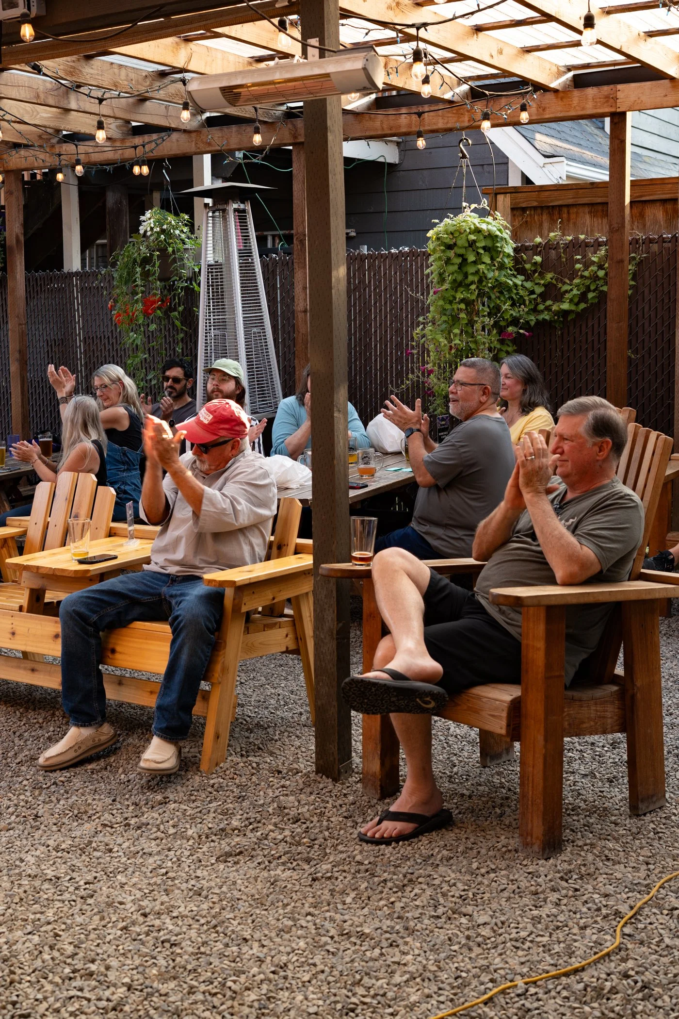 People sitting at outdoor wooden tables and chairs under a wooden pergola with string lights, enjoying drinks and socializing in a backyard setting.