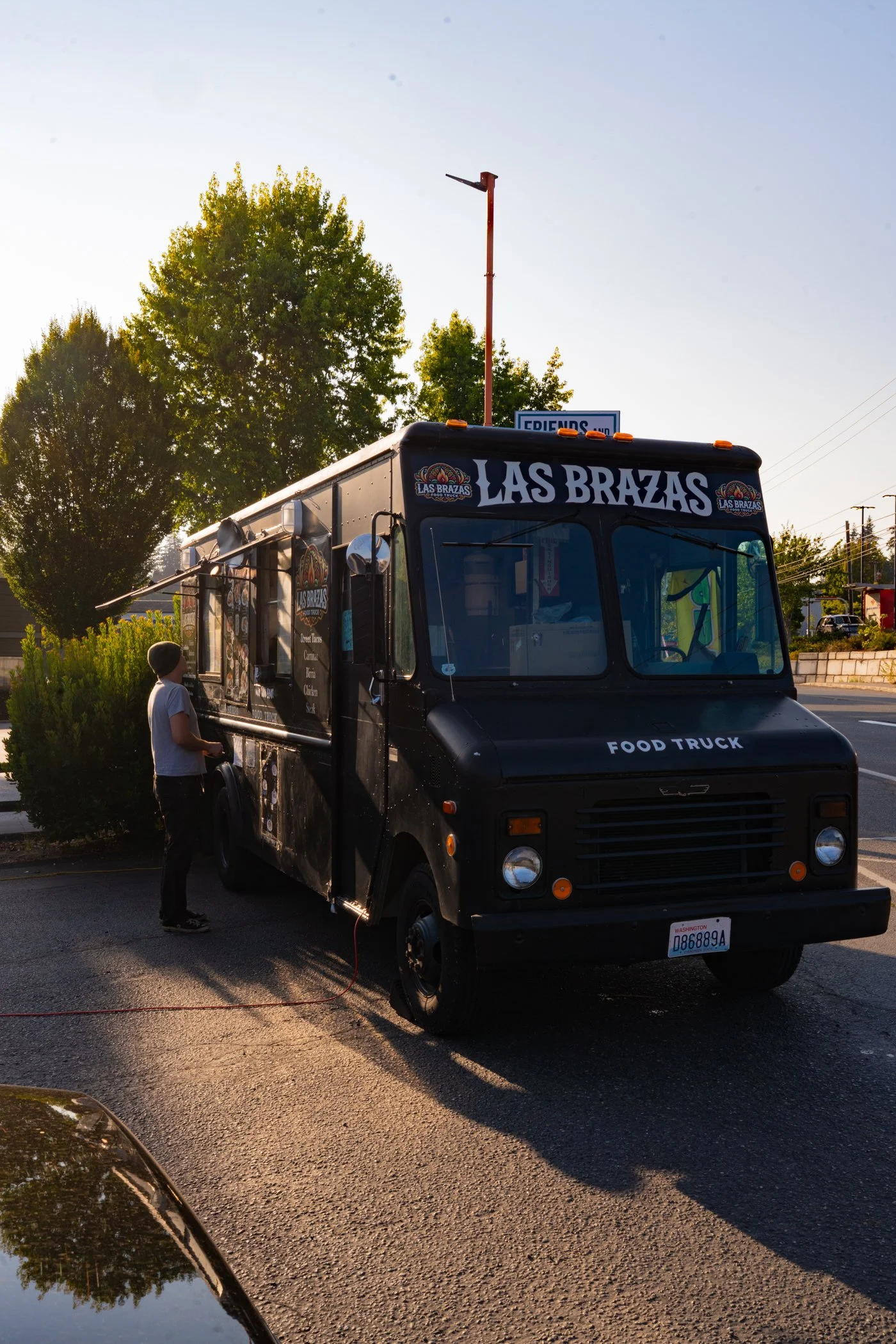 A black food truck named Las Brazas is parked on the side of the street during sunset. A person wearing a gray beanie and white shirt is standing near the truck. Tall trees and a street sign are visible in the background.