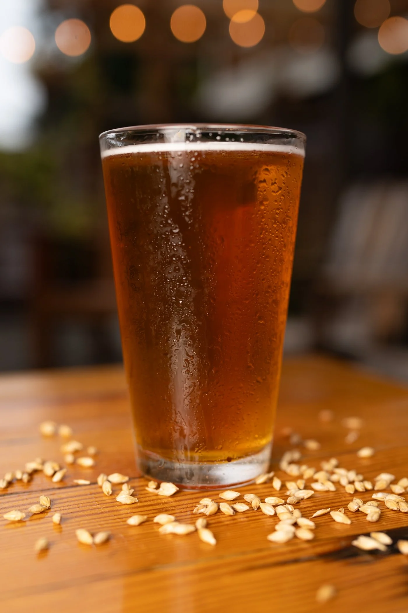 A glass of beer on a wooden surface with scattered oats around it, with a blurred background