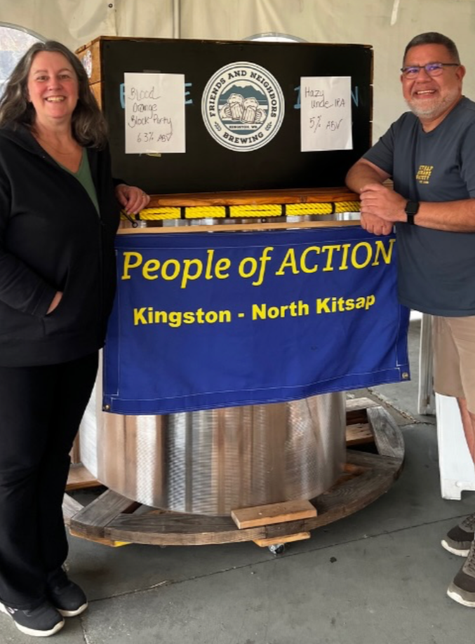 Two smiling people standing next to a large brew kettle with a banner that reads "People of ACTION Kingston - North Kitsap". The brew kettle has a box on top with notes indicating a blood orange block party and Hazy India Pale Ale.