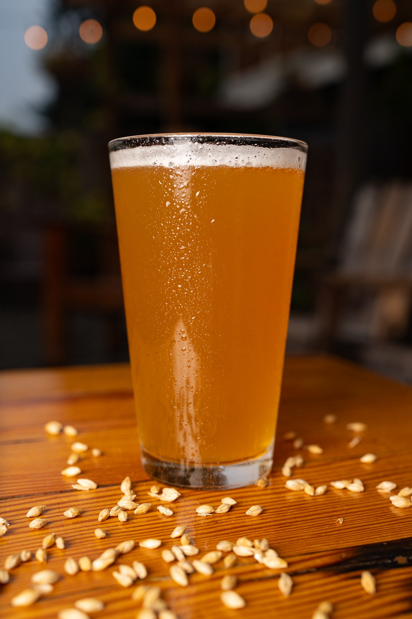 A tall glass filled with a golden, slightly cloudy beverage, topped with a thin layer of foam, sitting on a wooden surface with scattered oat flakes. In the background, blurred warm lights are visible.