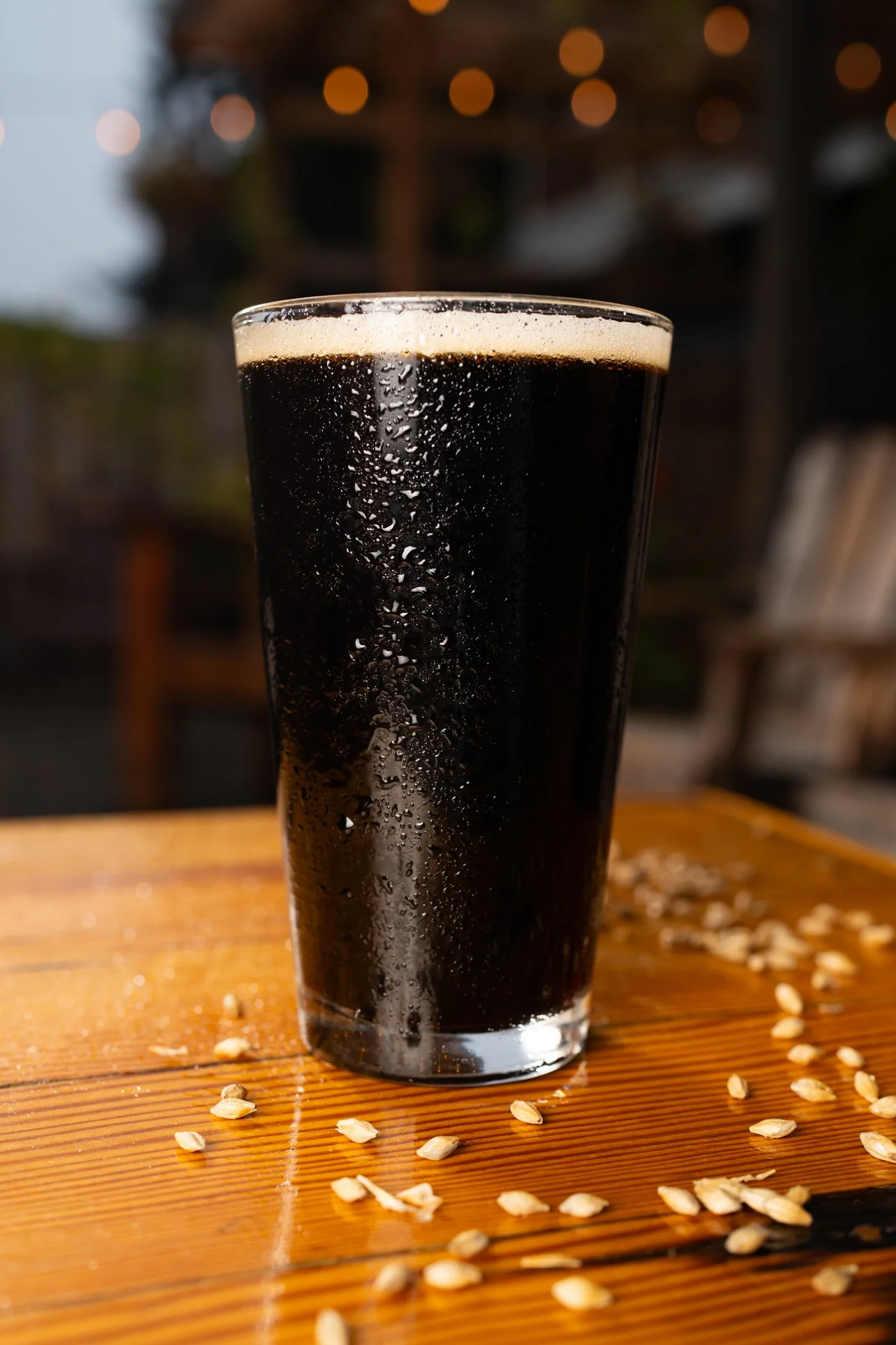 A glass of dark cola soda with condensation, placed on a wooden surface with scattered grain oats, background blurred with bokeh lights.