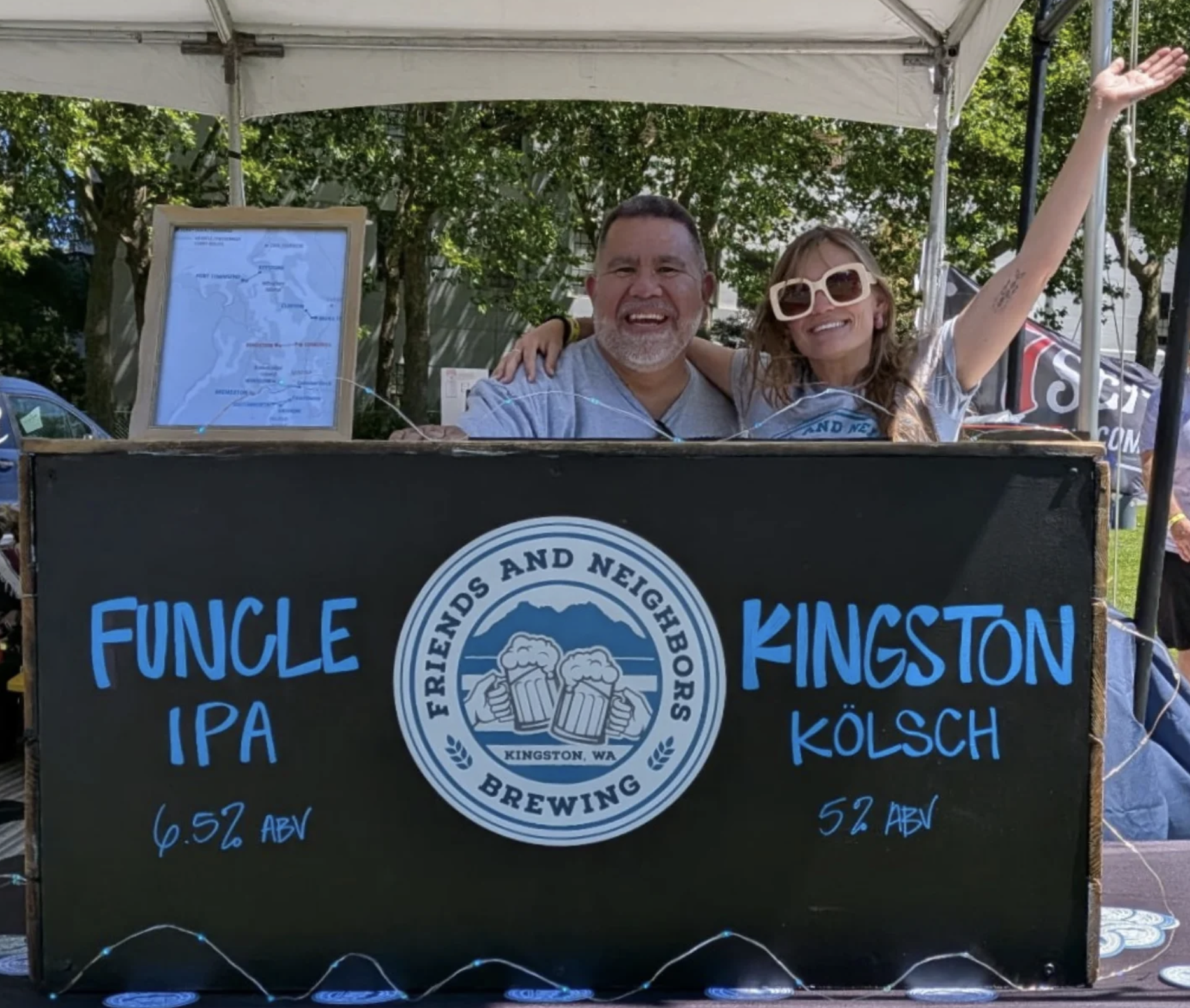A smiling man and woman at a beer stand outdoors, with trees and a tent in the background. The woman's arm is raised, and they are standing behind a black banner with blue and white text advertising Kingston Brewing, featuring a logo with two clinking beer mugs.