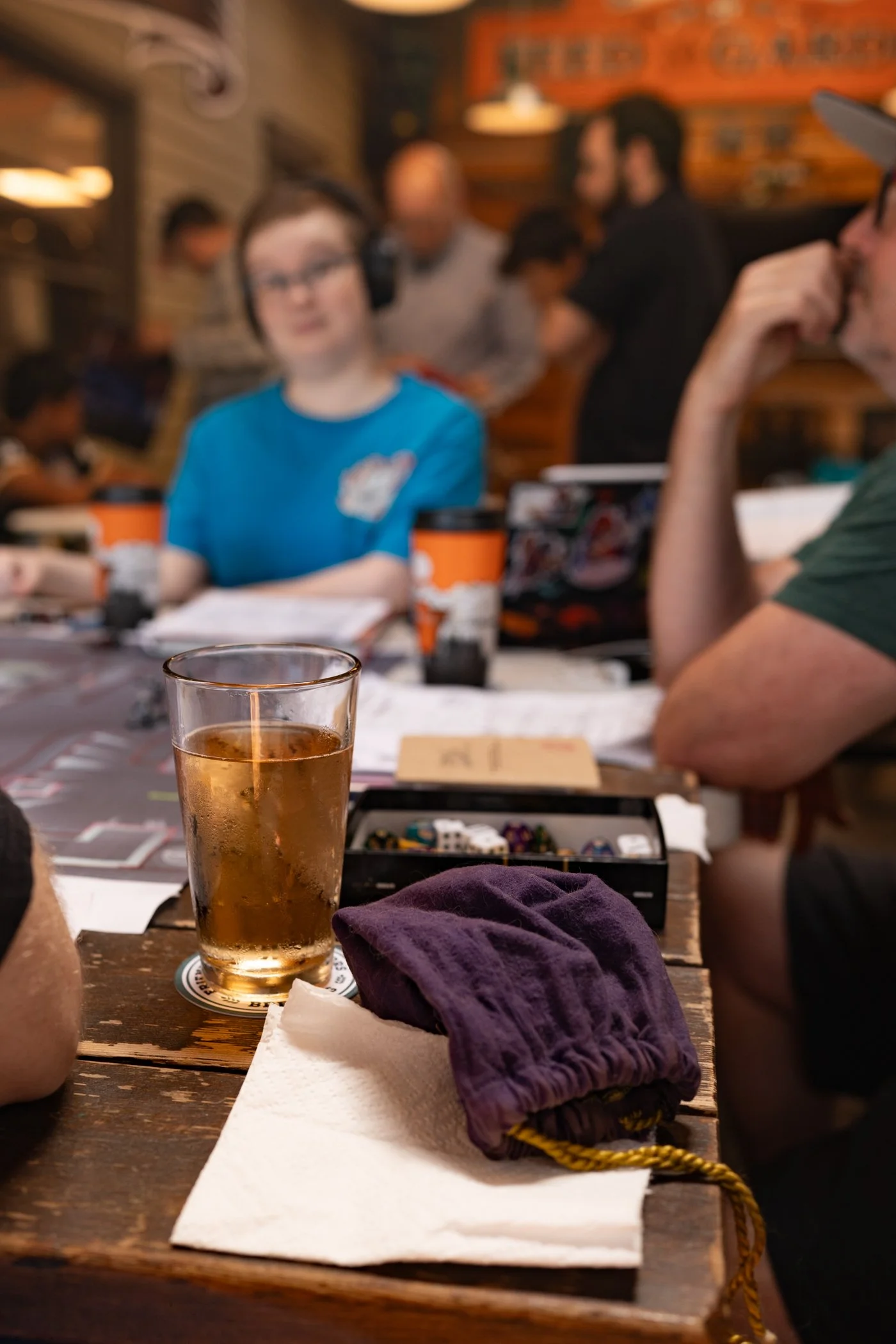 A glass of beer on a wooden table with a purple cloth, napkin, and a box of gaming dice in the background. Several people are in the background of a rustic cafe or bar setting, engaged in playing a board game, with some wearing headphones.