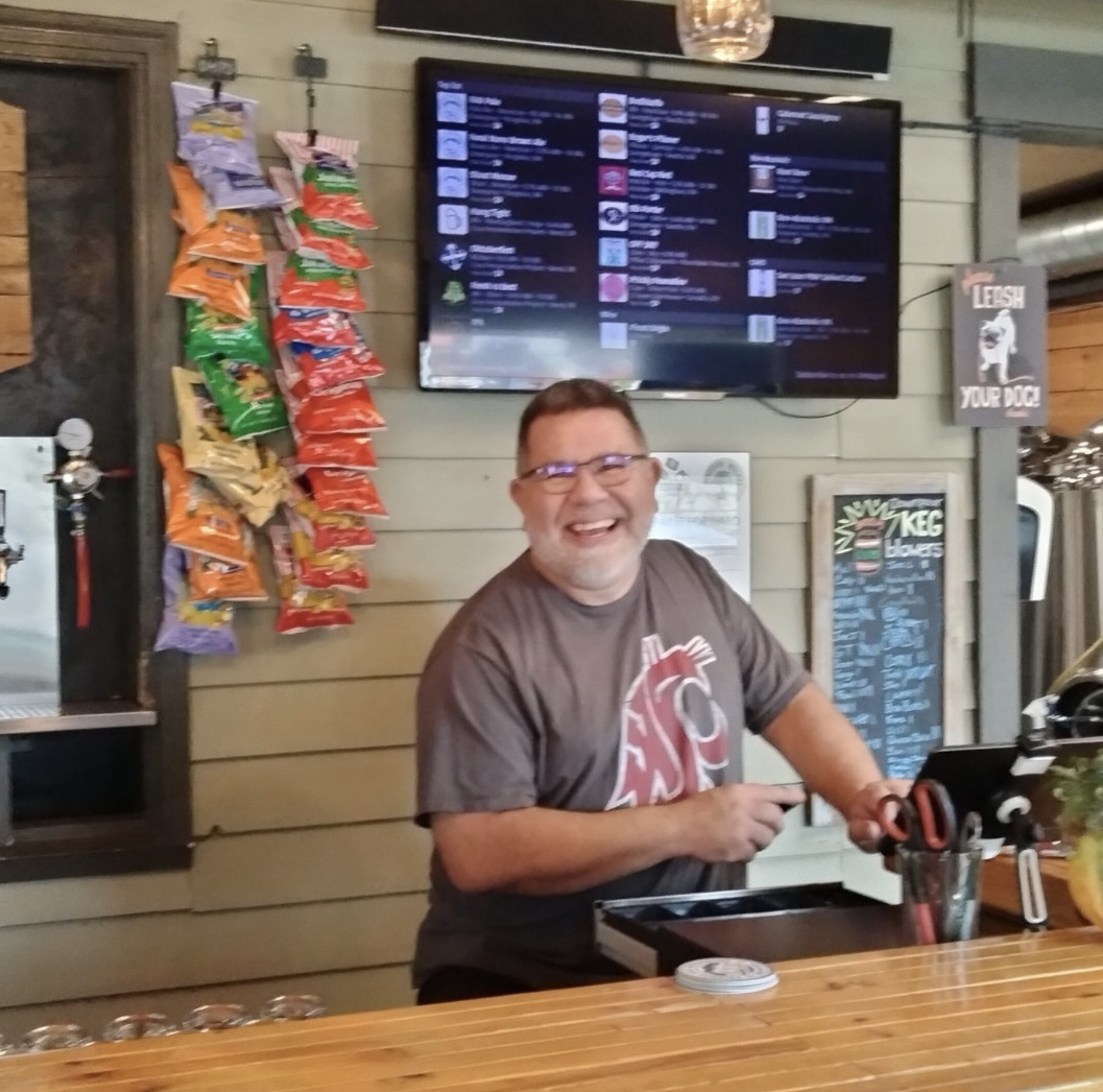 A smiling man with glasses wearing a gray t-shirt with red and white lettering, sitting at a bar in a restaurant or pub, holding a mobile phone. Behind him is a wall-mounted menu screen, a decorative hanging light, and a display of snack bags hanging on a rack to the left.
