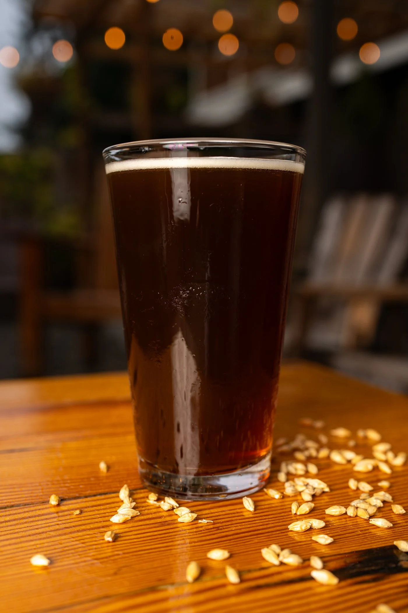A tall glass of dark beer with a foamy head, placed on a wooden table sprinkled with scattered oats. The background is blurred with warm bokeh lights.
