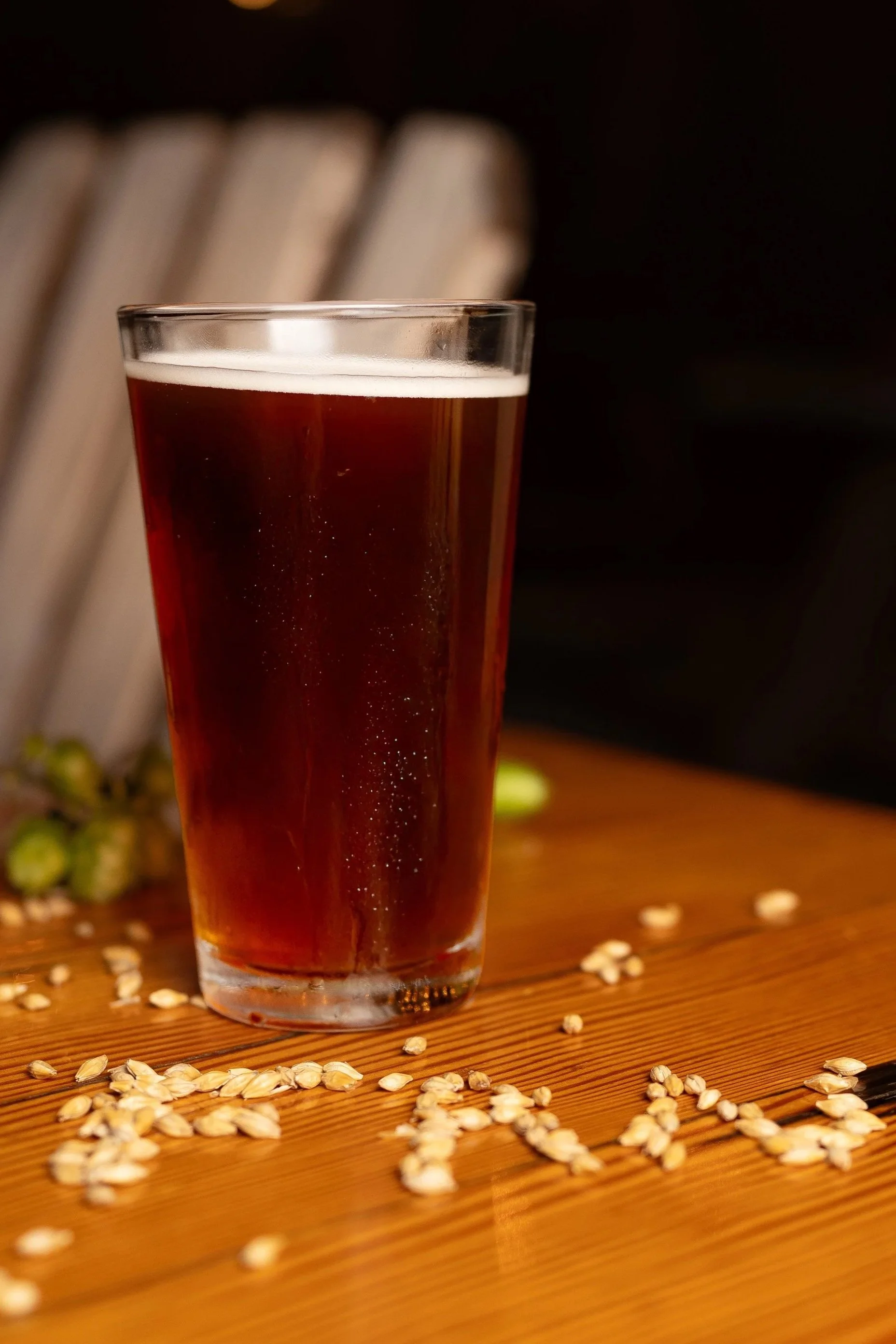 A glass of dark beer on a wooden surface with scattered oats and a blurred background.