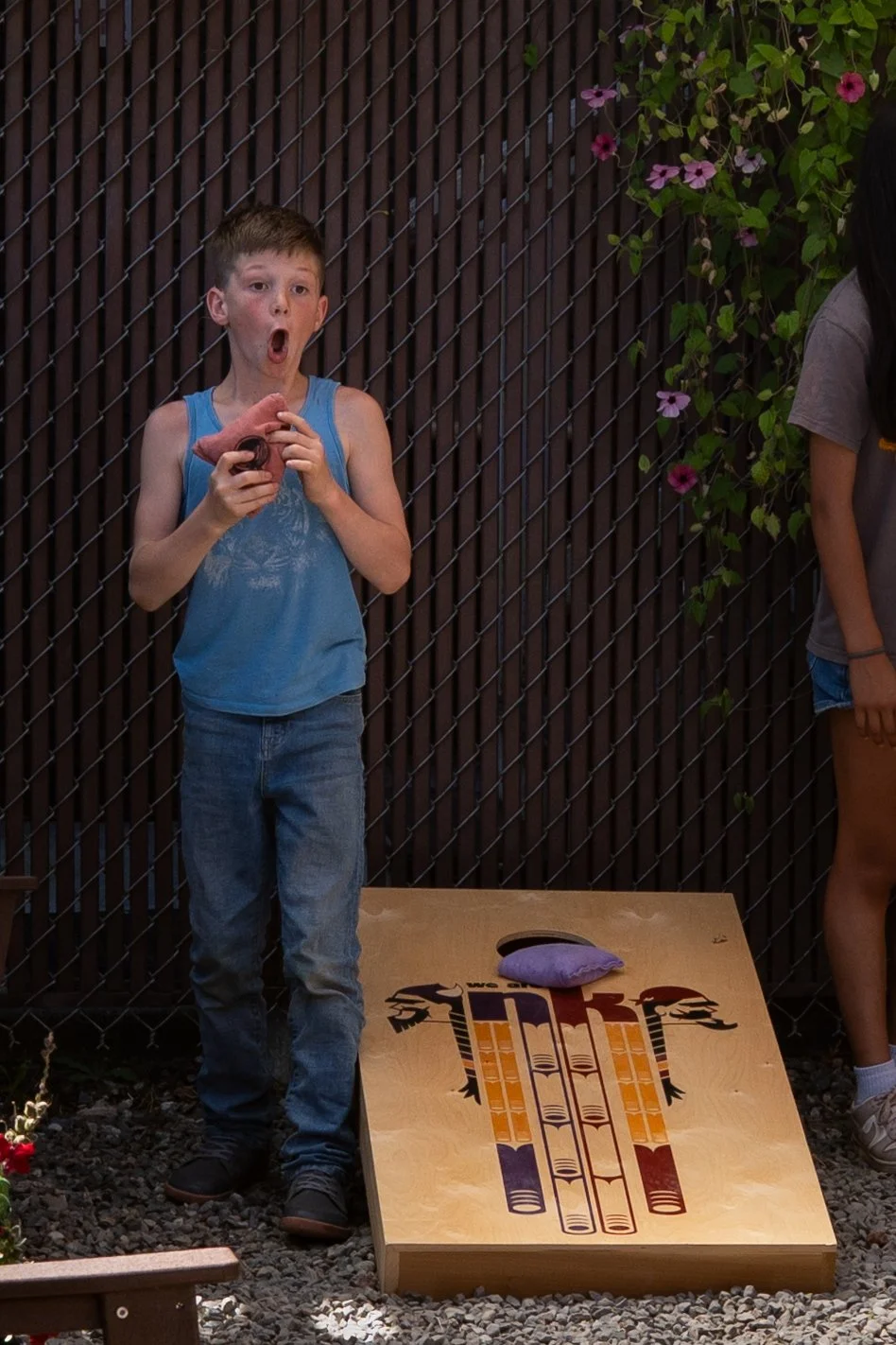 A young boy in a blue tank top and jeans playing cornhole, holding a bean bag with his mouth open, in an outdoor setting with a wooden fence and flowers in the background.