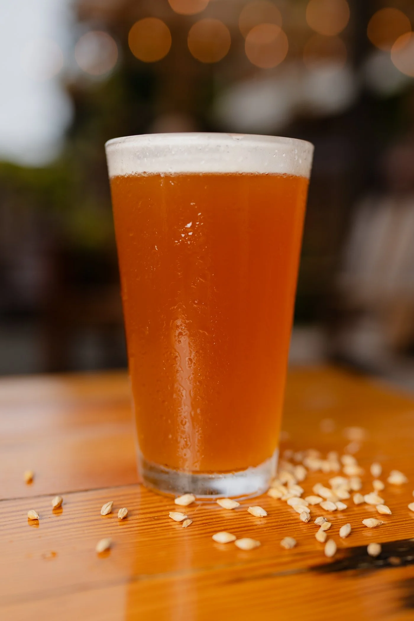 A glass of beer on a wooden surface with scattered oats around it and a blurred background with warm bokeh lights.