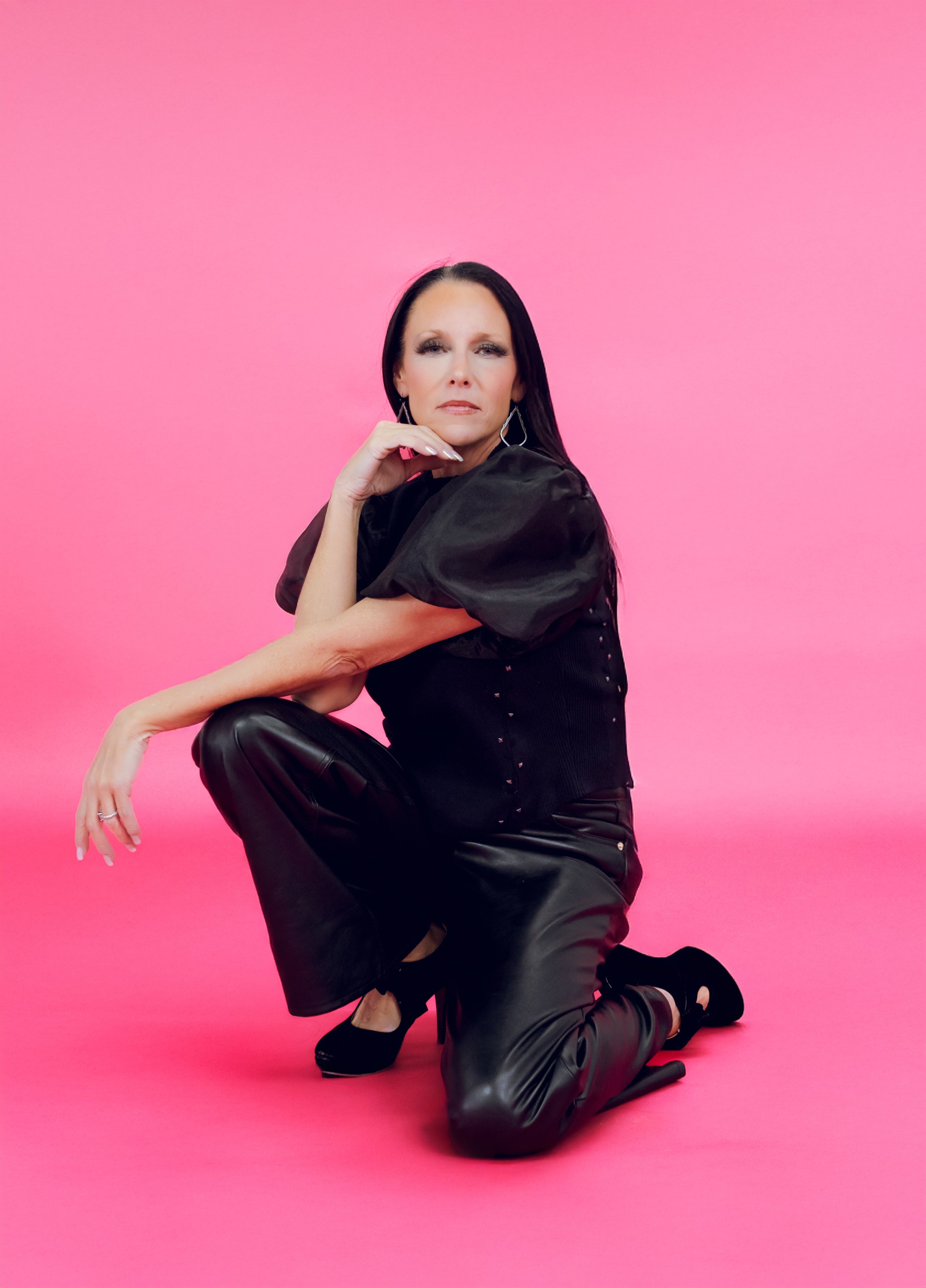 Woman with black hair, wearing black outfit with puffed sleeves and black shoes, kneeling with one knee on pink background, resting her hand on her chin, looking at the camera.