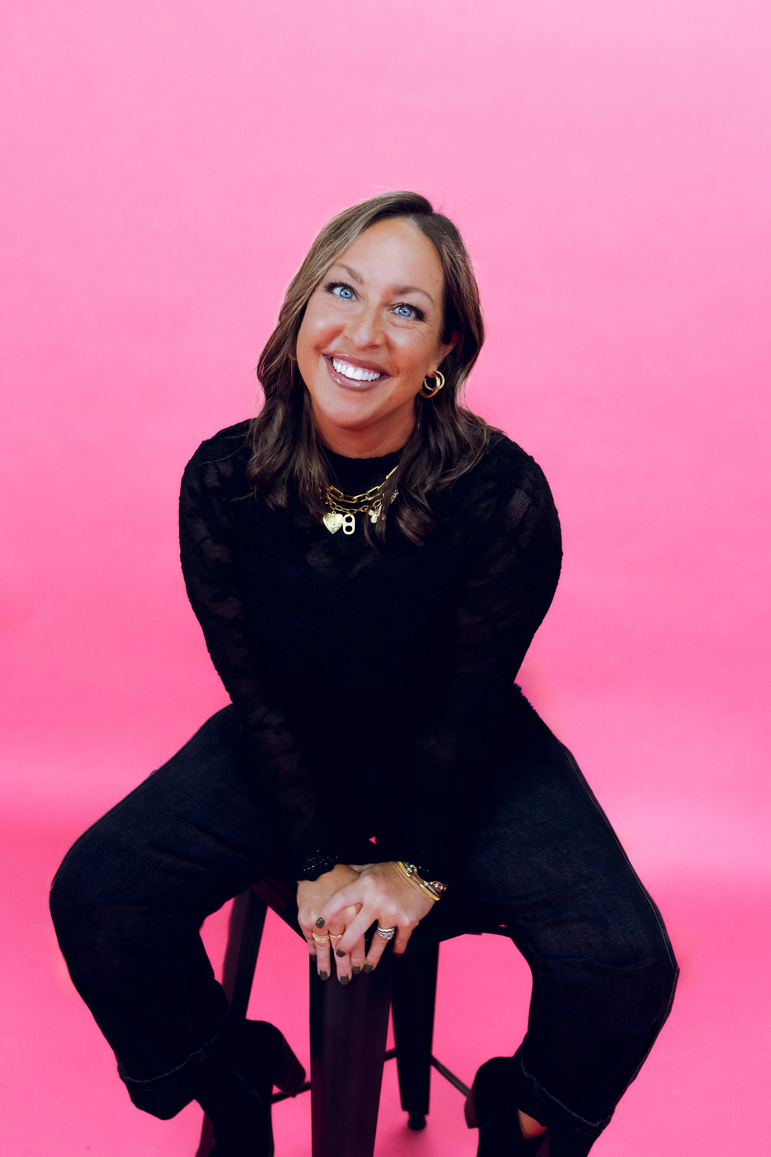Woman sitting on a black stool against a pink background, smiling and looking at the camera. She has shoulder-length brown hair and is wearing a black long-sleeve top, layered gold necklaces, gold hoop earrings, rings, and bracelets.