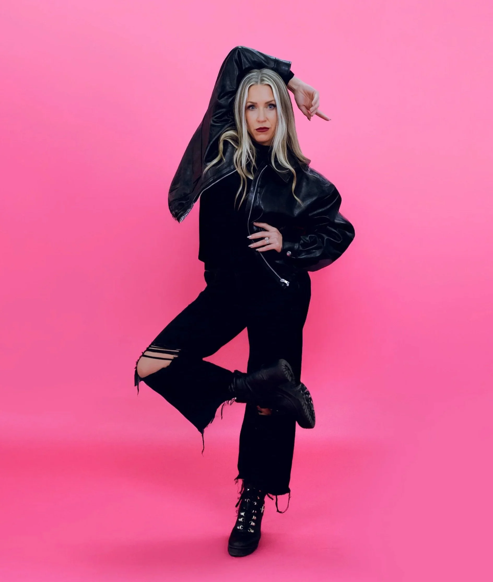 A woman with long blonde hair wearing a black leather jacket, black ripped jeans, and black lace-up boots, striking a dance pose against a pink background.