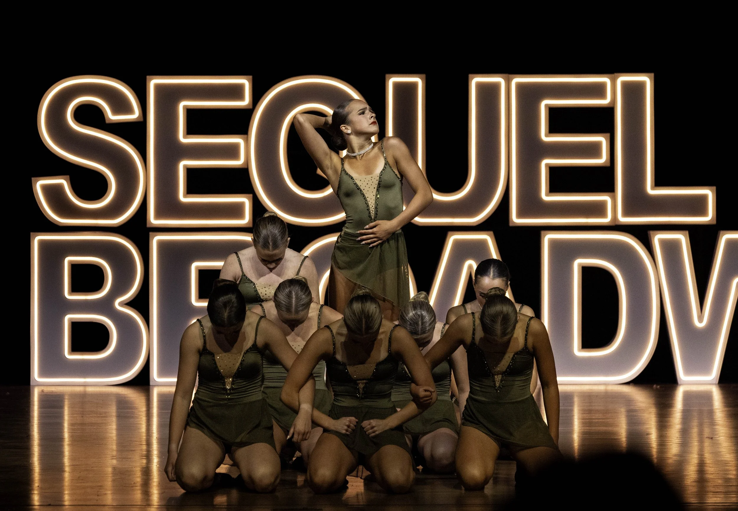 A group of women in matching dance costumes performing on stage with a large illuminated 'SEQUEL BROADWAY' sign in the background.