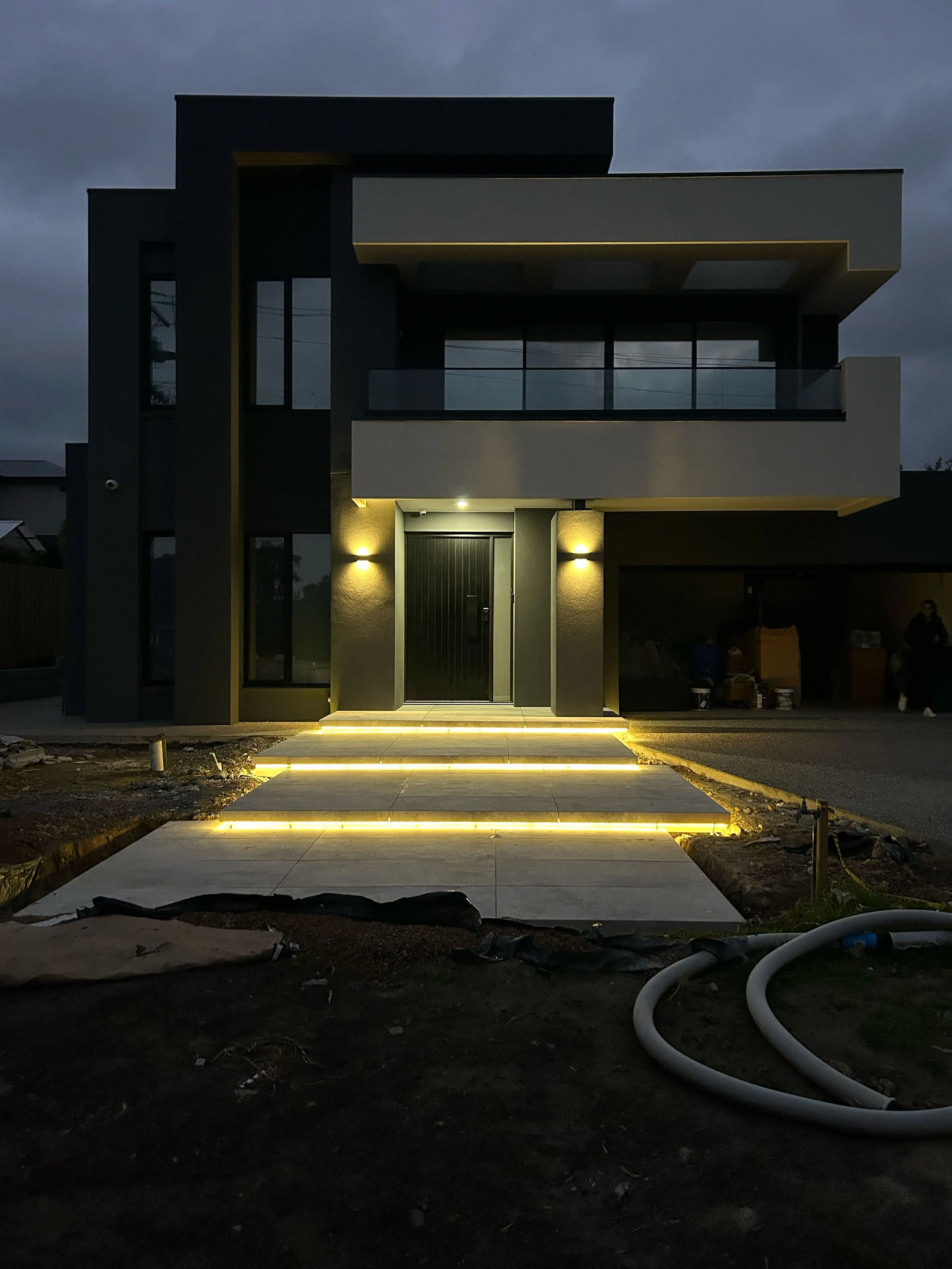 Modern two-story house with lighting along stairs and around entrance, dark exterior, large balcony, cloudy evening sky.