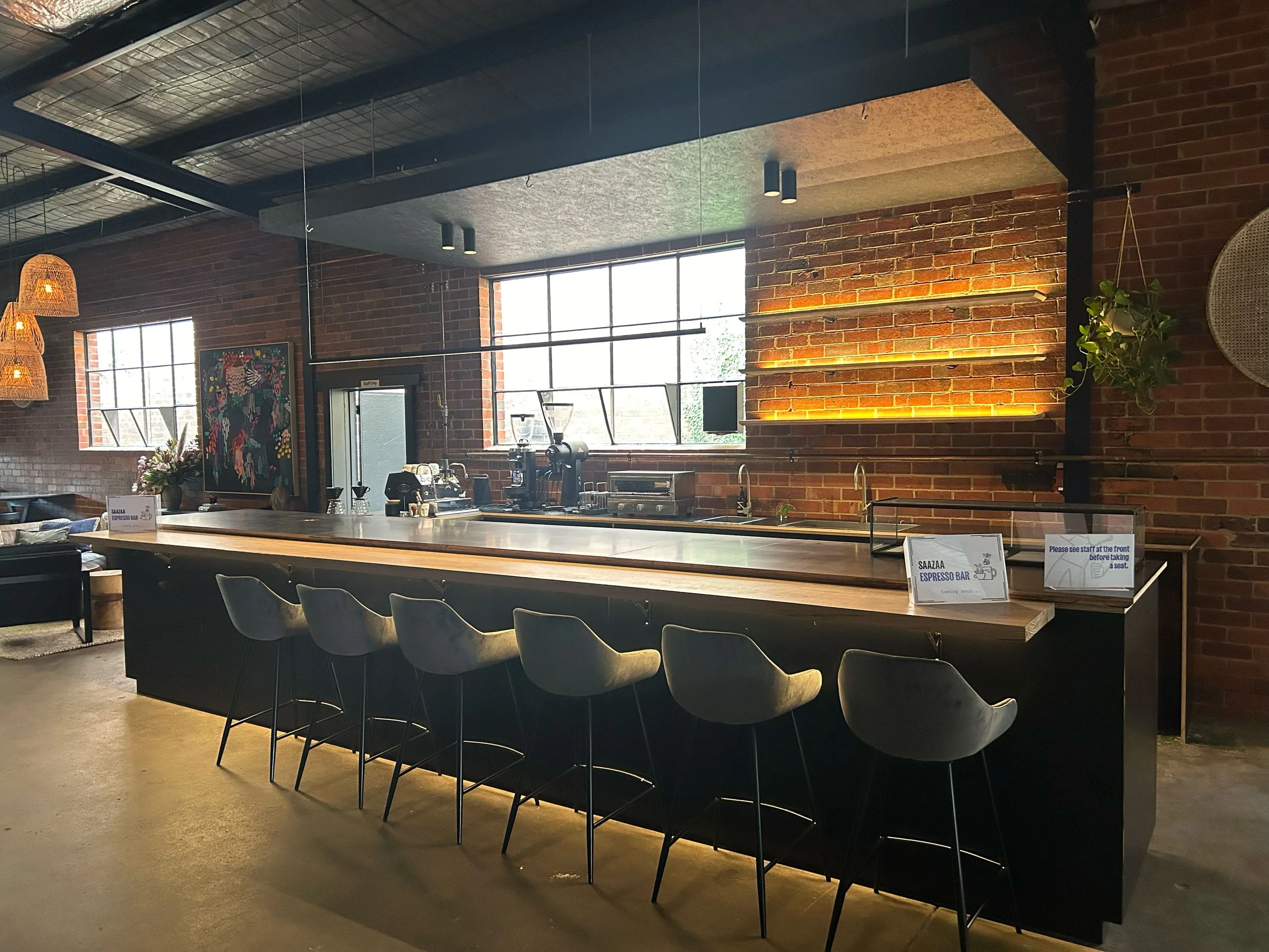 Modern coffee shop interior with brick walls, large windows, a long wooden counter with six chairs, and cozy seating area in the background.
