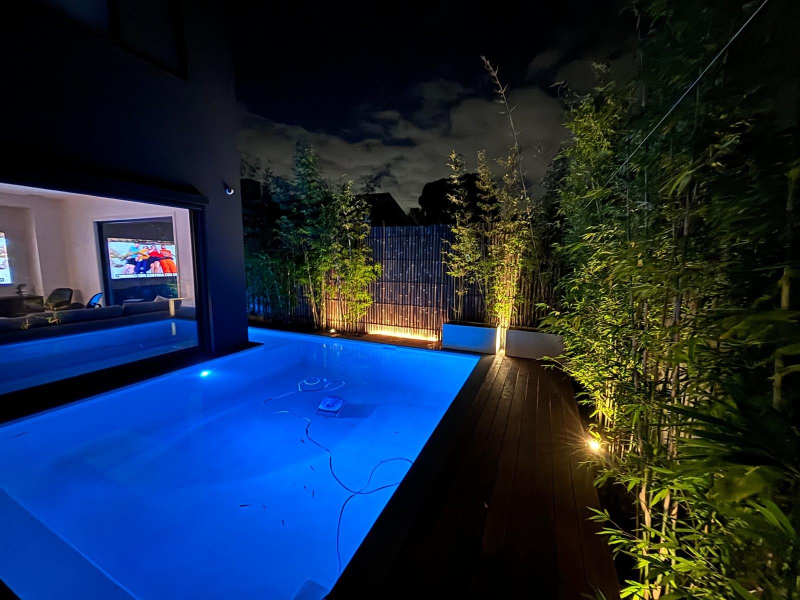 Night view of an outdoor pool illuminated with blue lights, surrounded by greenery and wooden decking, with a modern house interior visible through large windows and a cloudy sky above.