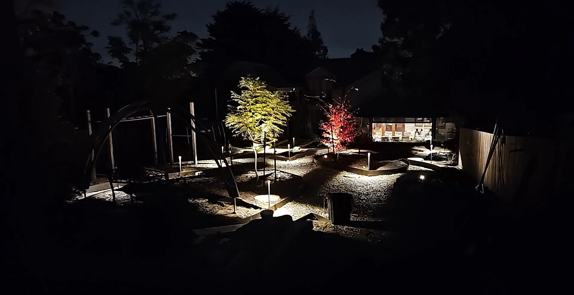 Nighttime view of a backyard garden with illuminated trees, garden beds, a shed, and a fence, featuring decorative outdoor lighting.