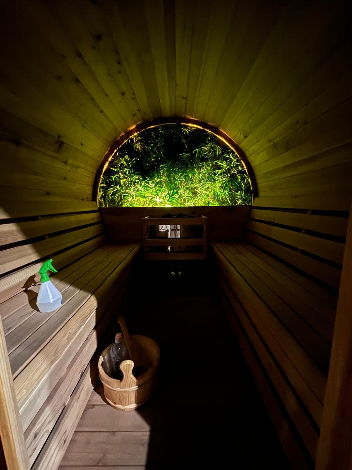 Wood-paneled interior of a sauna with a window revealing lush green plants outside, a spray bottle on a wooden bench, and a bucket with a ladle inside.