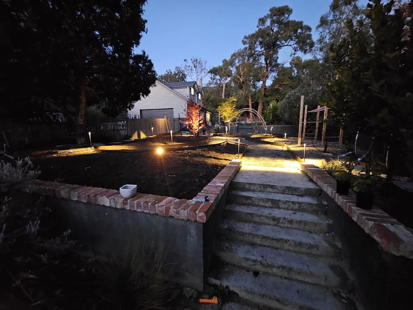 Nighttime view of a backyard with steps leading up to a garden, illuminated by outdoor lights, with trees, plants, and a house in the background.