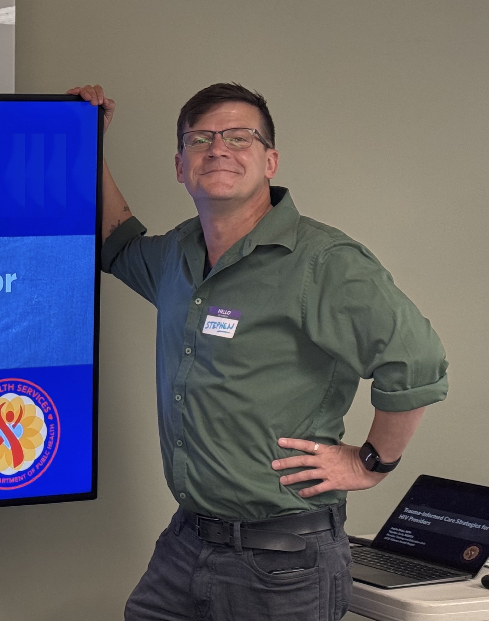 A man standing indoors next to a large monitor, wearing glasses, a green shirt, and a name tag that reads 'Stephen'. He is smiling and has his left hand on his hip.