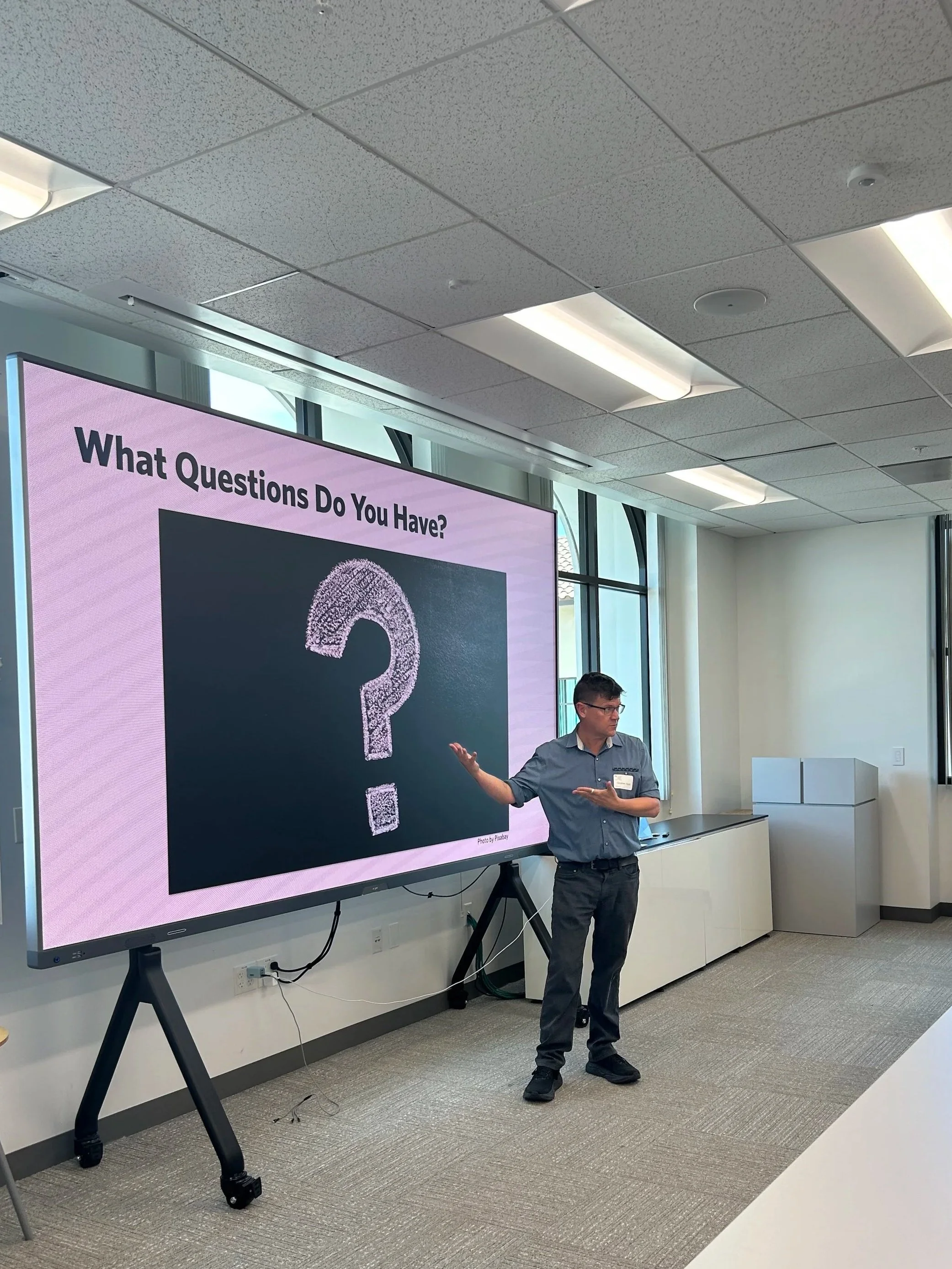 A man giving a presentation in a conference room with a large screen displaying the text 'What Questions Do You Have?' and a large question mark.