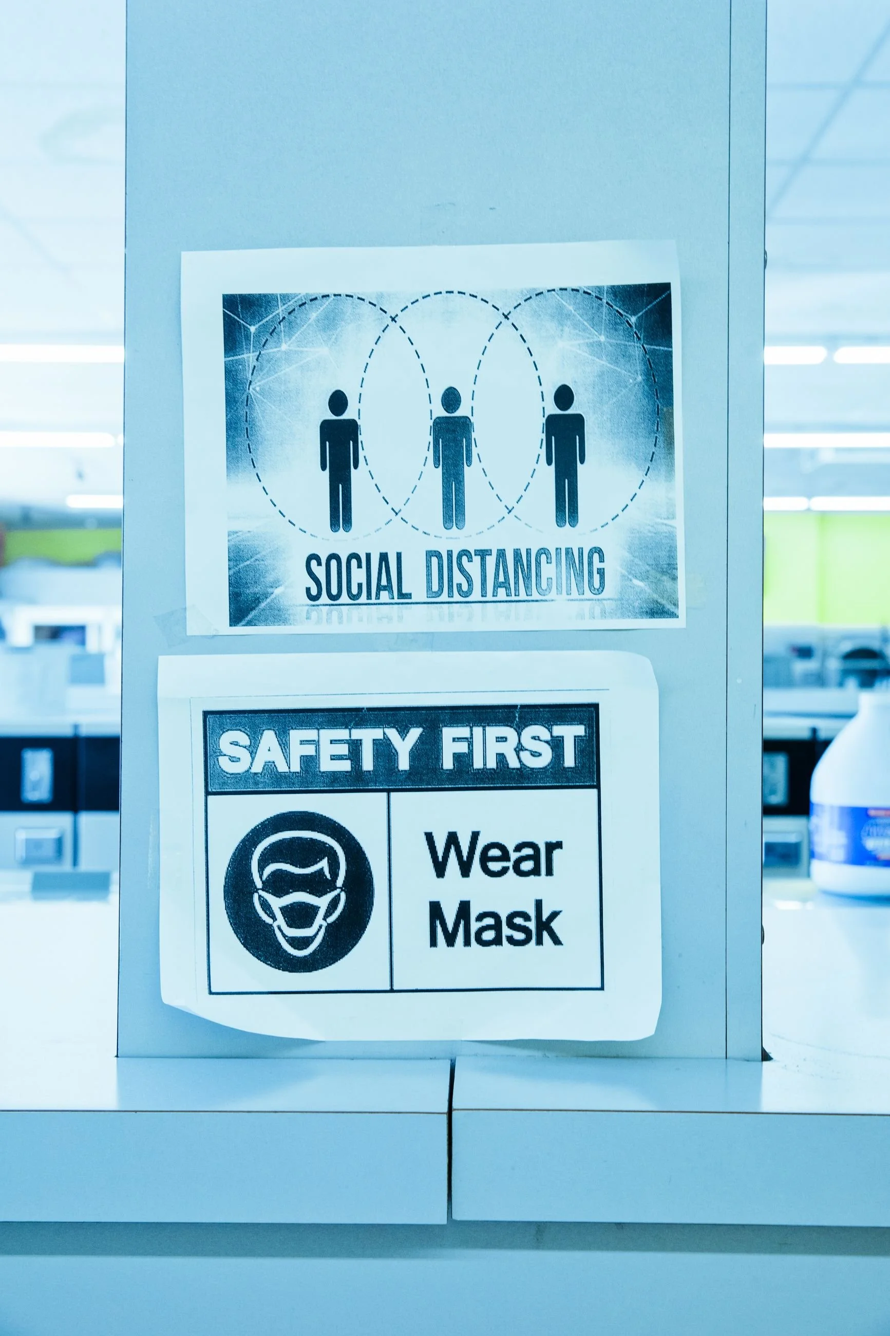 A sign on a wall in a public space with safety instructions for social distancing and wearing masks, including illustrations of people standing apart and wearing face masks.