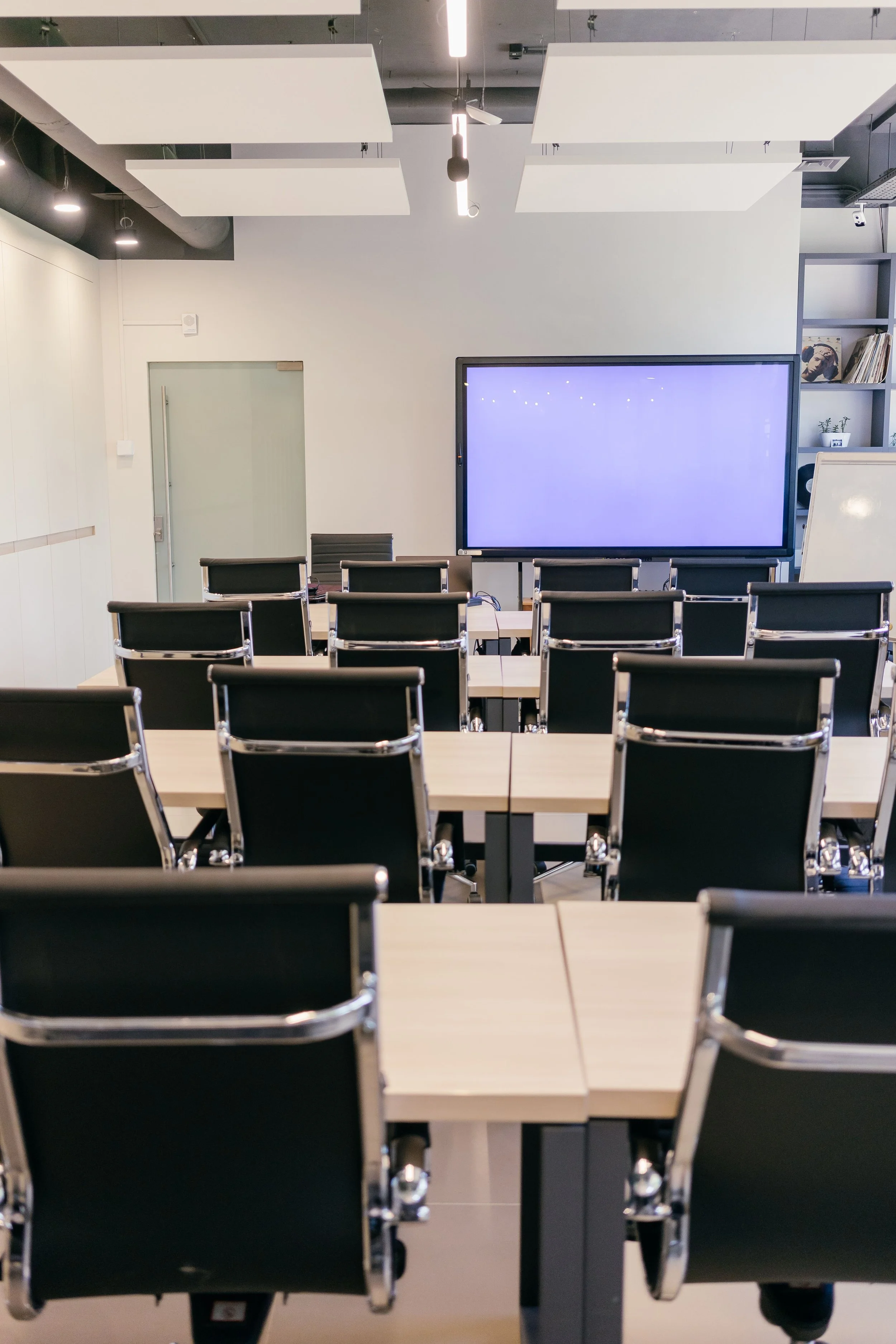 A modern conference room with flexible seating, a large digital screen at the front, and a whiteboard on the side, in a well-lit space with ceiling sound panels.